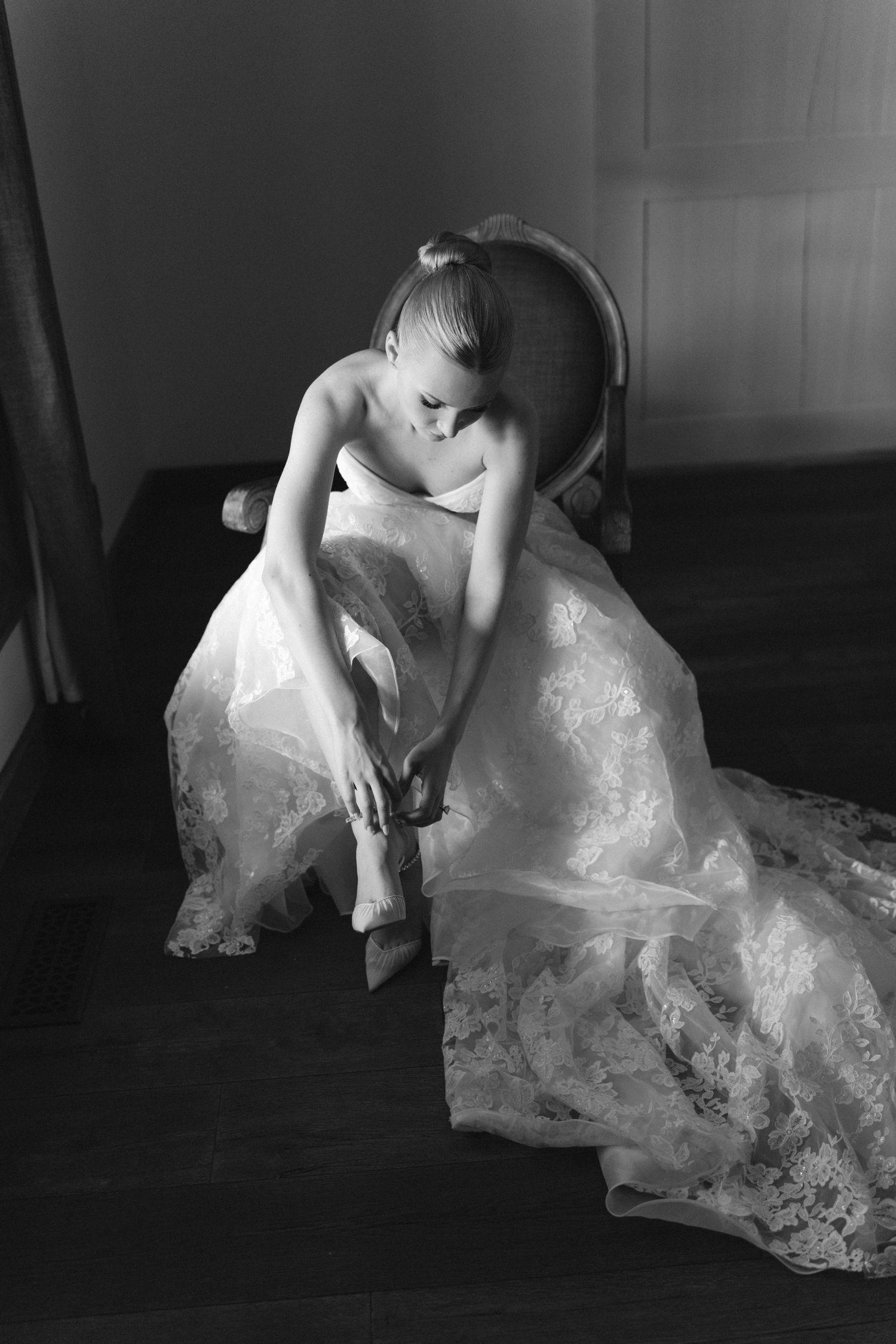Black and white photo of a bride in a lace gown sitting on a chair putting on her wedding shoes.