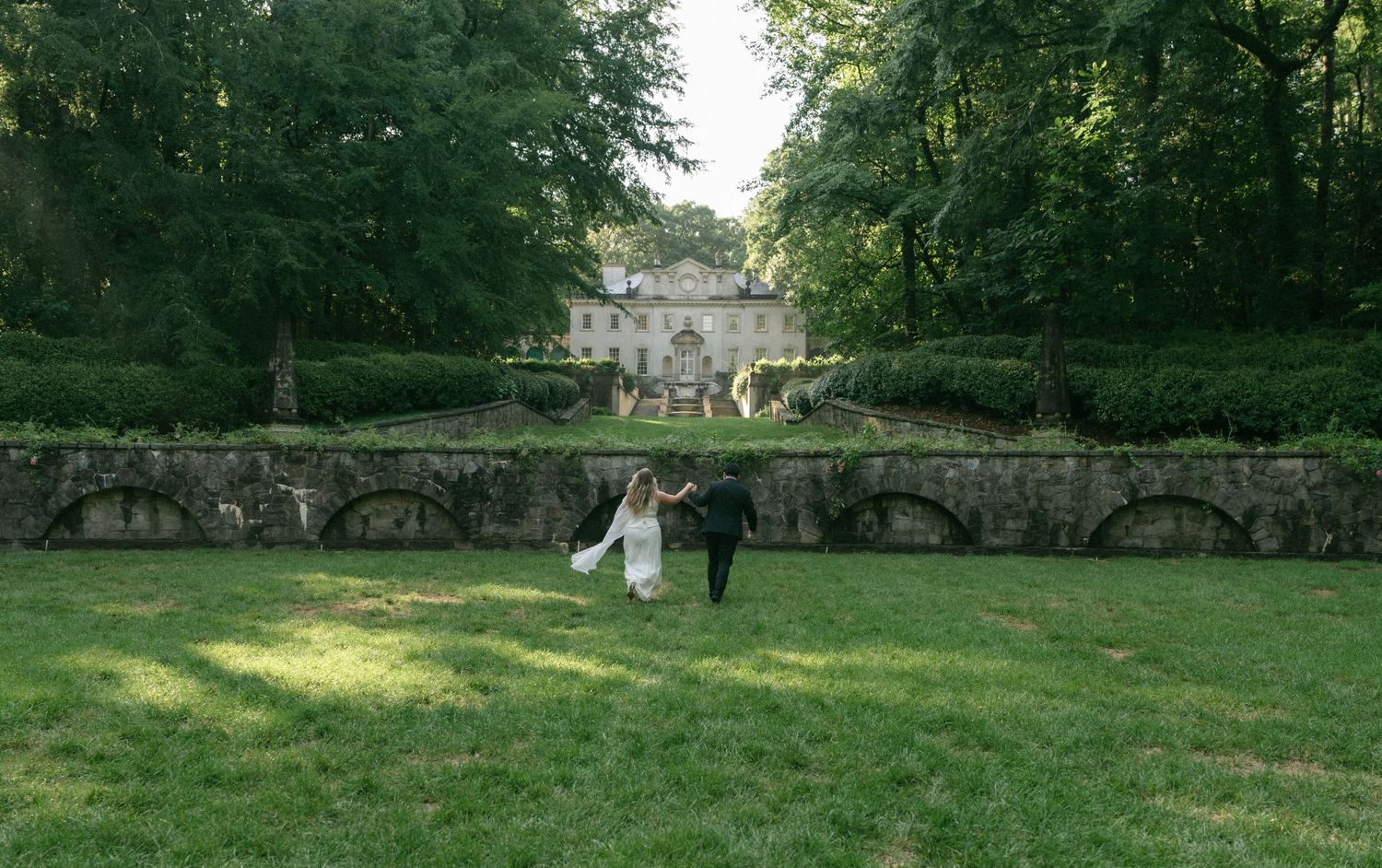 Bride and groom walking hand in hand toward an elegant stone mansion surrounded by lush green gardens.