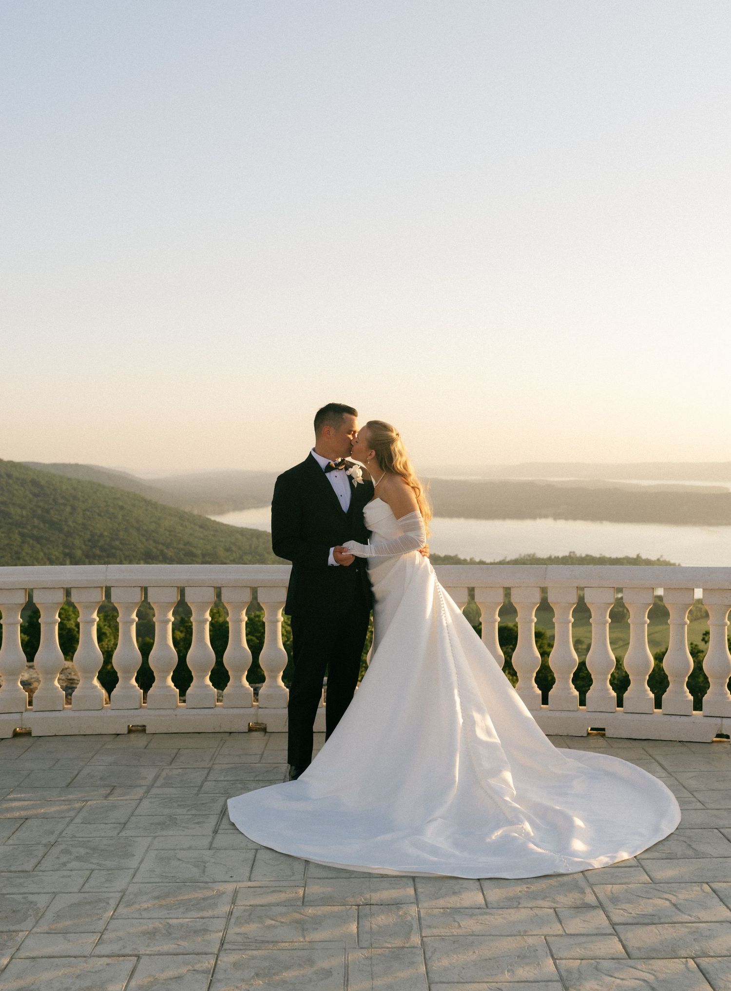 Bride and groom share a romantic kiss on a stone terrace overlooking a scenic lake and rolling hills at sunset.