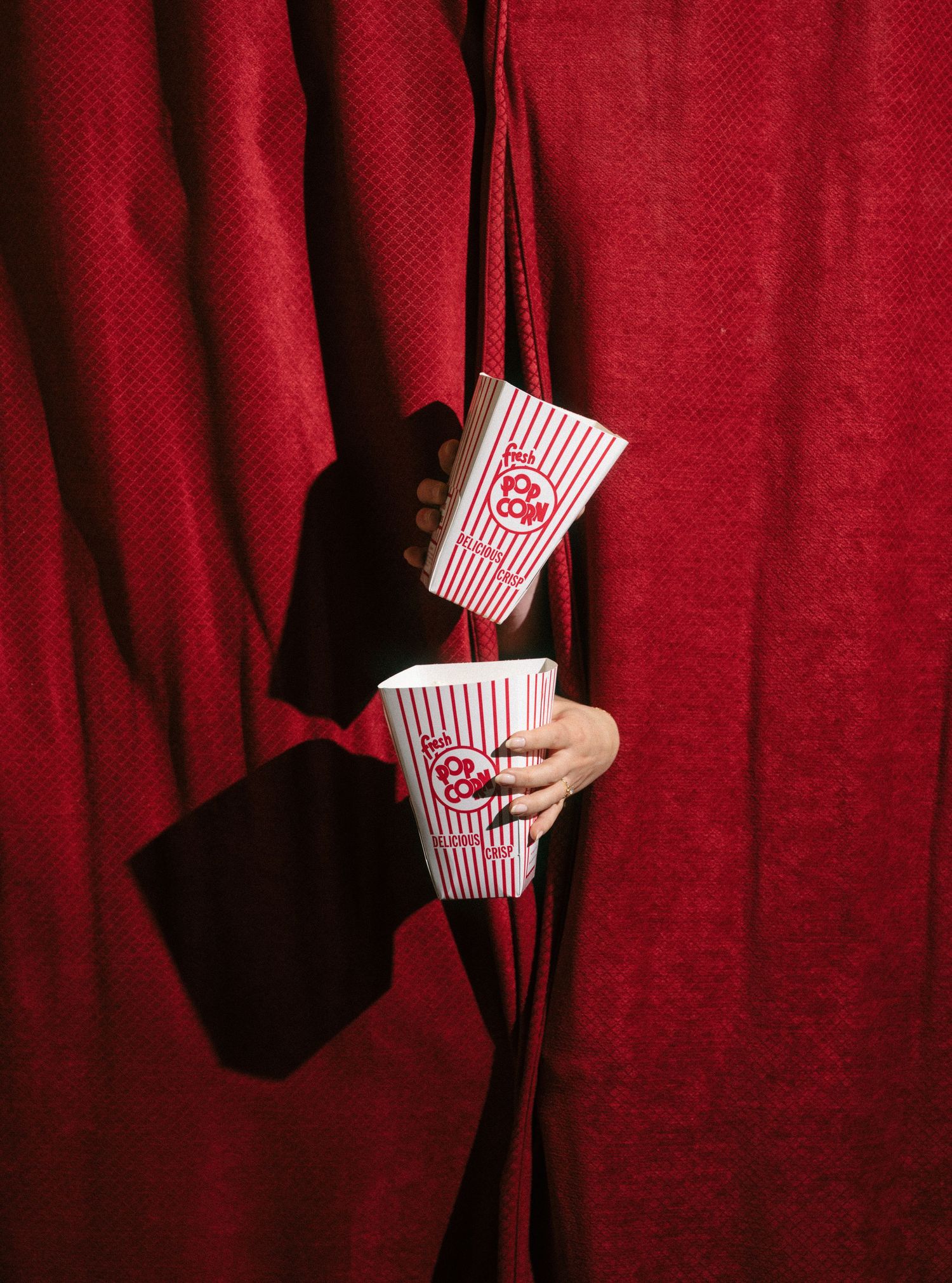 Two hands emerging from red curtains holding striped popcorn boxes at a movie theater.