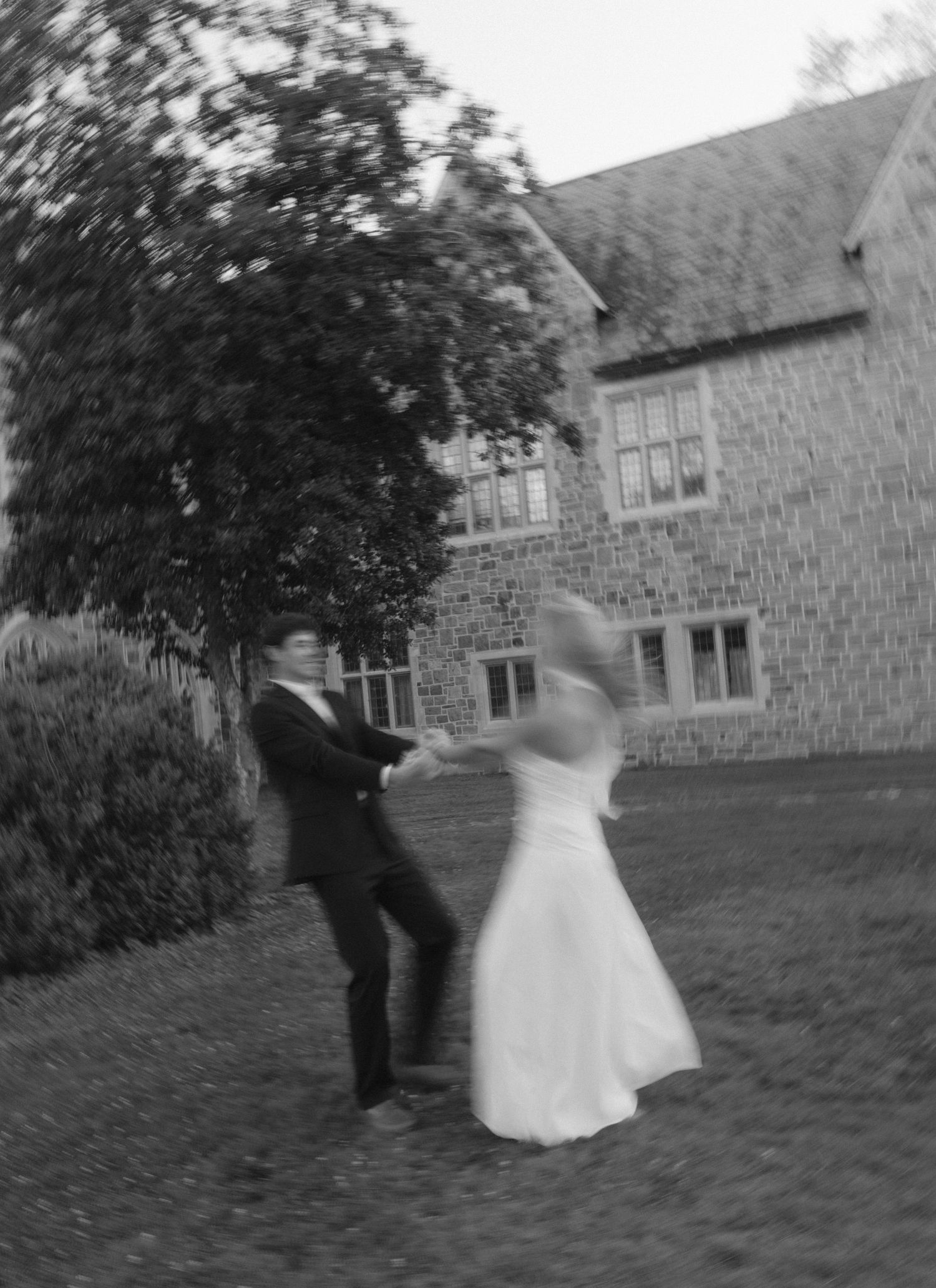 Black and white photo of a bride and groom dancing outdoors near a stone building, captured with motion blur.