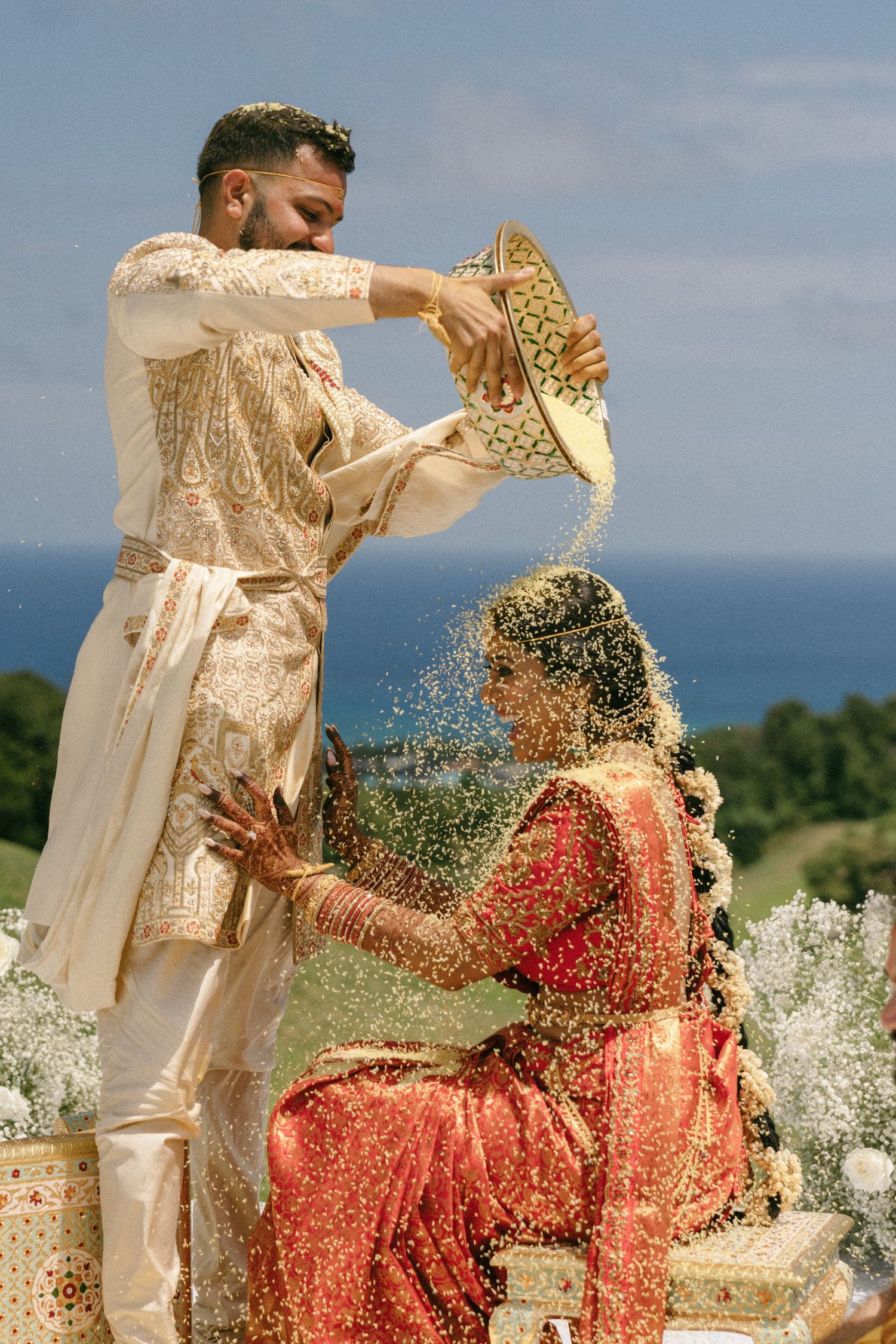 Groom pouring rice over bride during traditional Indian wedding ceremony outdoors with ocean backdrop.