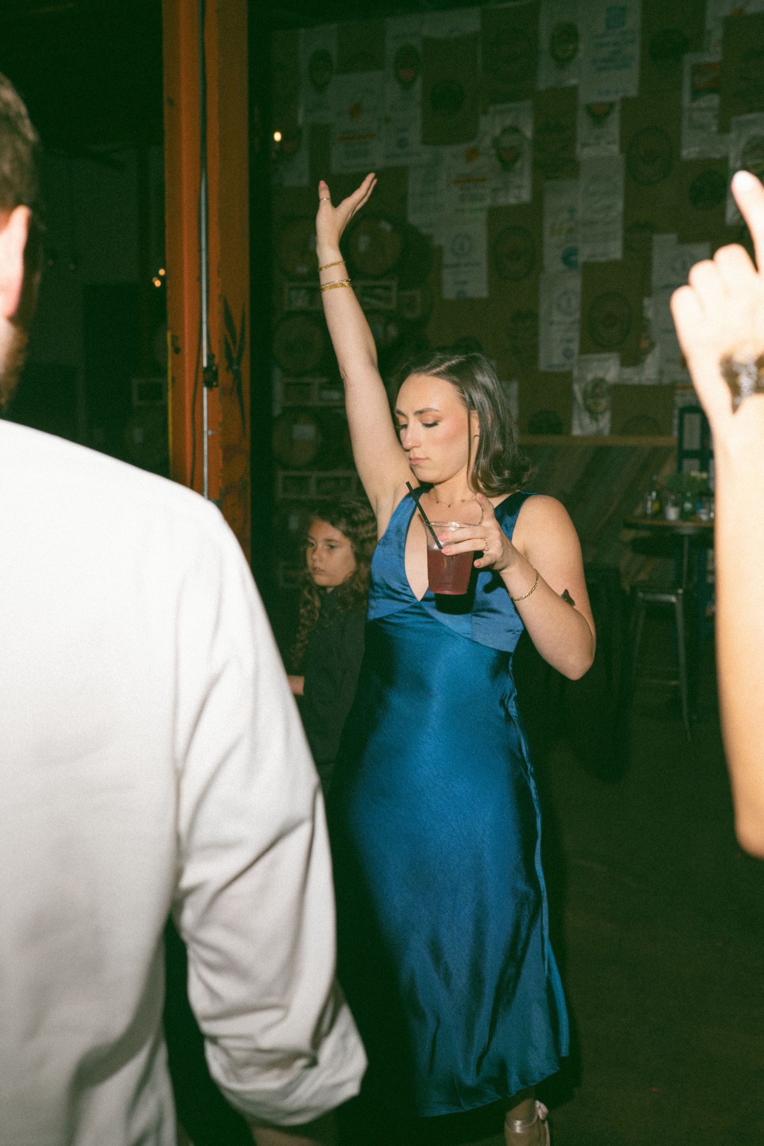 Woman in blue satin dress dancing with arm raised, holding a drink at an indoor party or event.