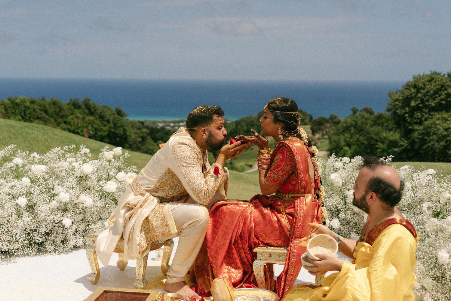 Couple in traditional Indian wedding attire sharing a romantic moment outdoors with lush greenery and ocean backdrop.