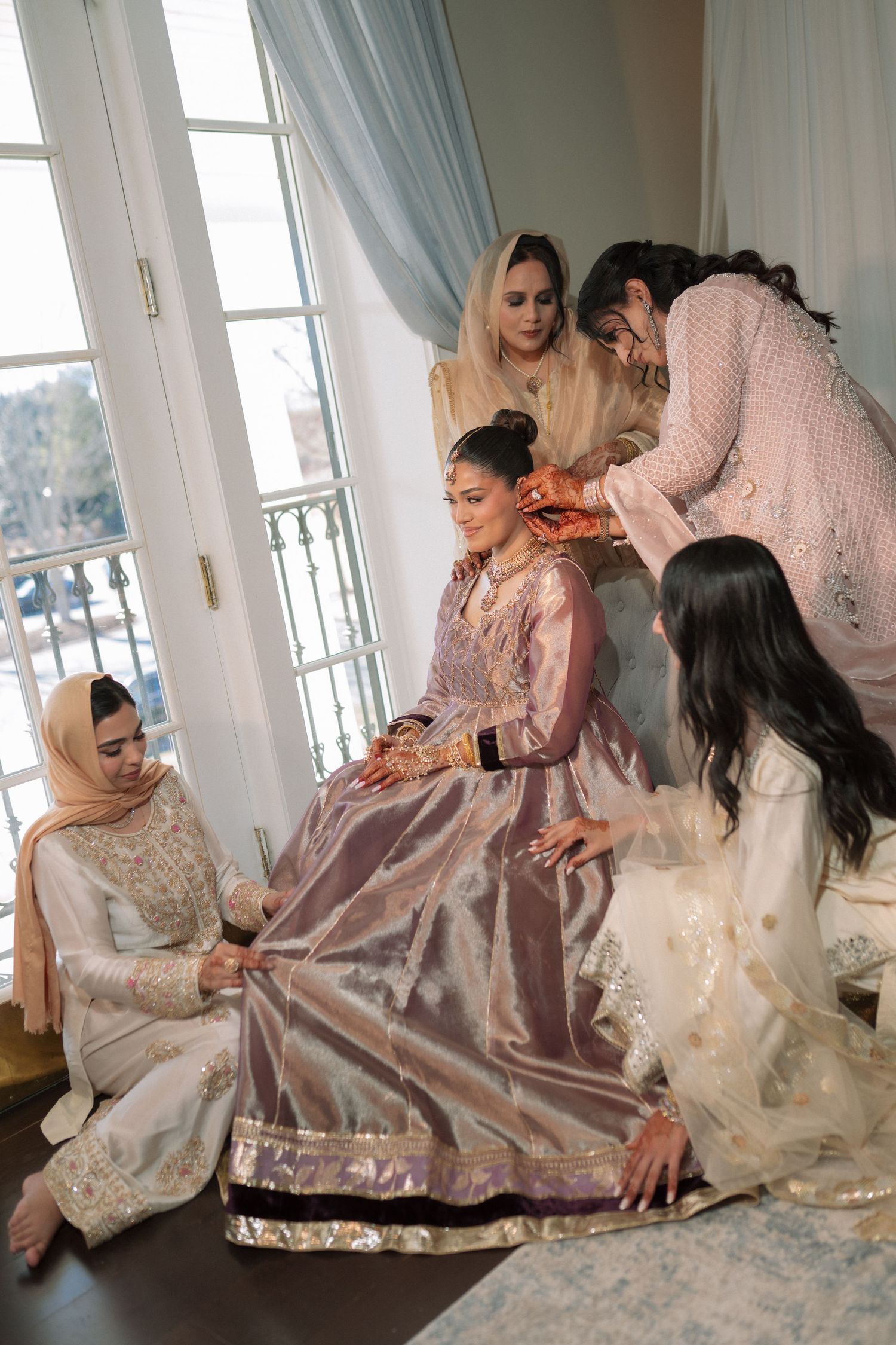 South Asian bride in lavender lehenga gets ready as bridesmaids in white traditional attire assist her near elegant windows.