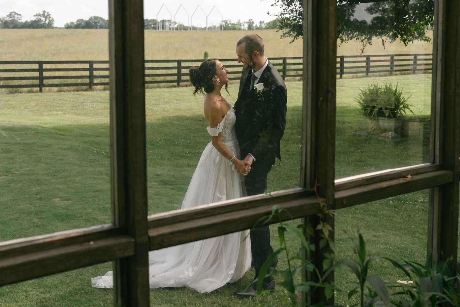 Bride and groom share a romantic moment outdoors, viewed through rustic windows with green fields and wooden fences behind them.