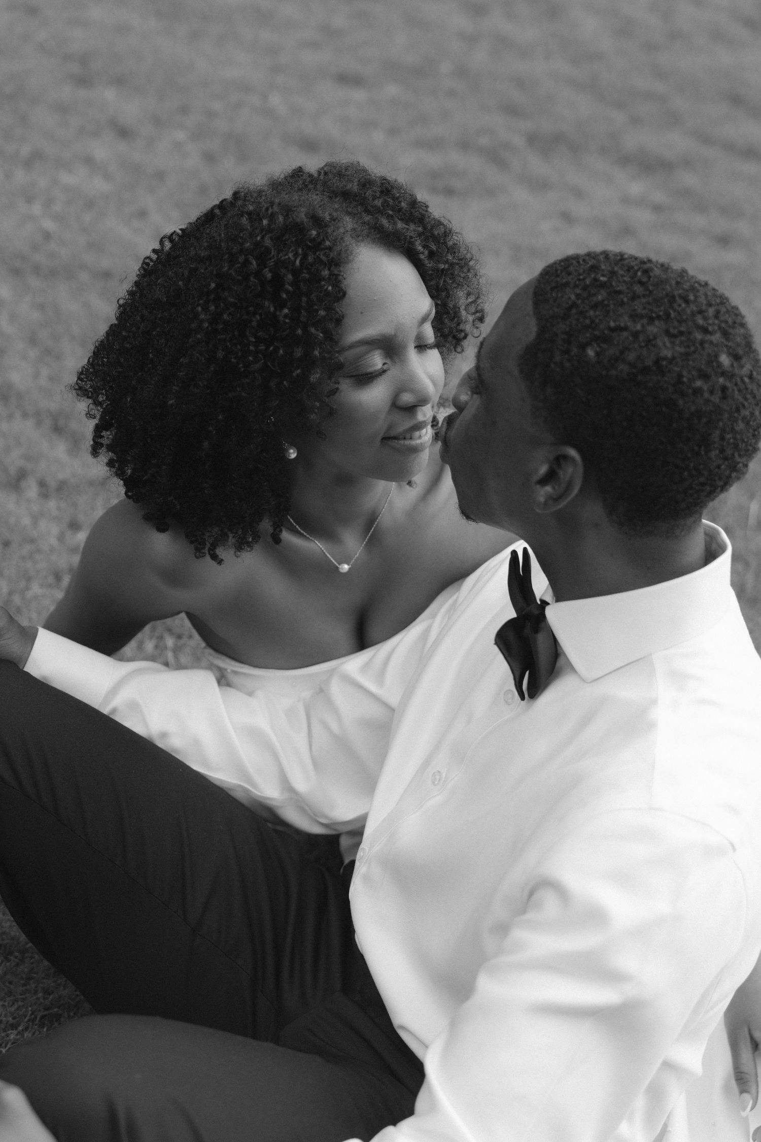 Black and white photo of a smiling bride and groom sitting together on grass.