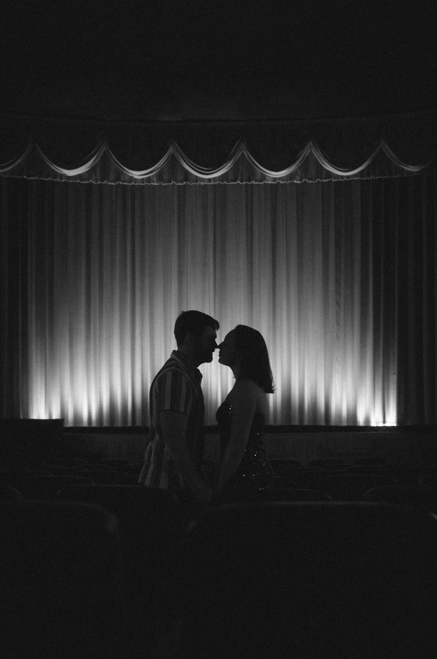 Black and white silhouette of a couple about to kiss in front of a glowing theater stage curtain.
