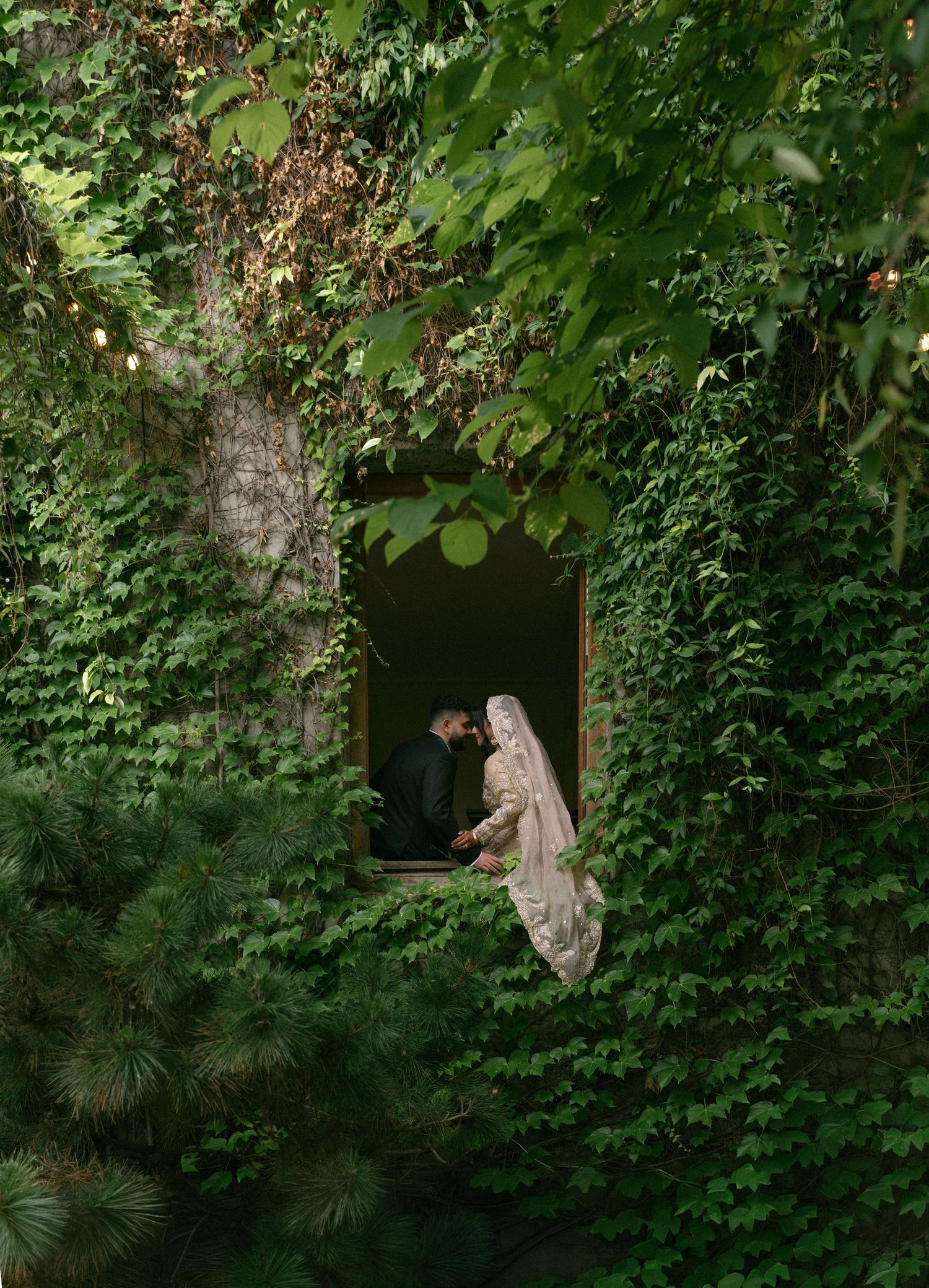 A bride and groom share an intimate moment in an ivy-covered stone doorway surrounded by lush green foliage.