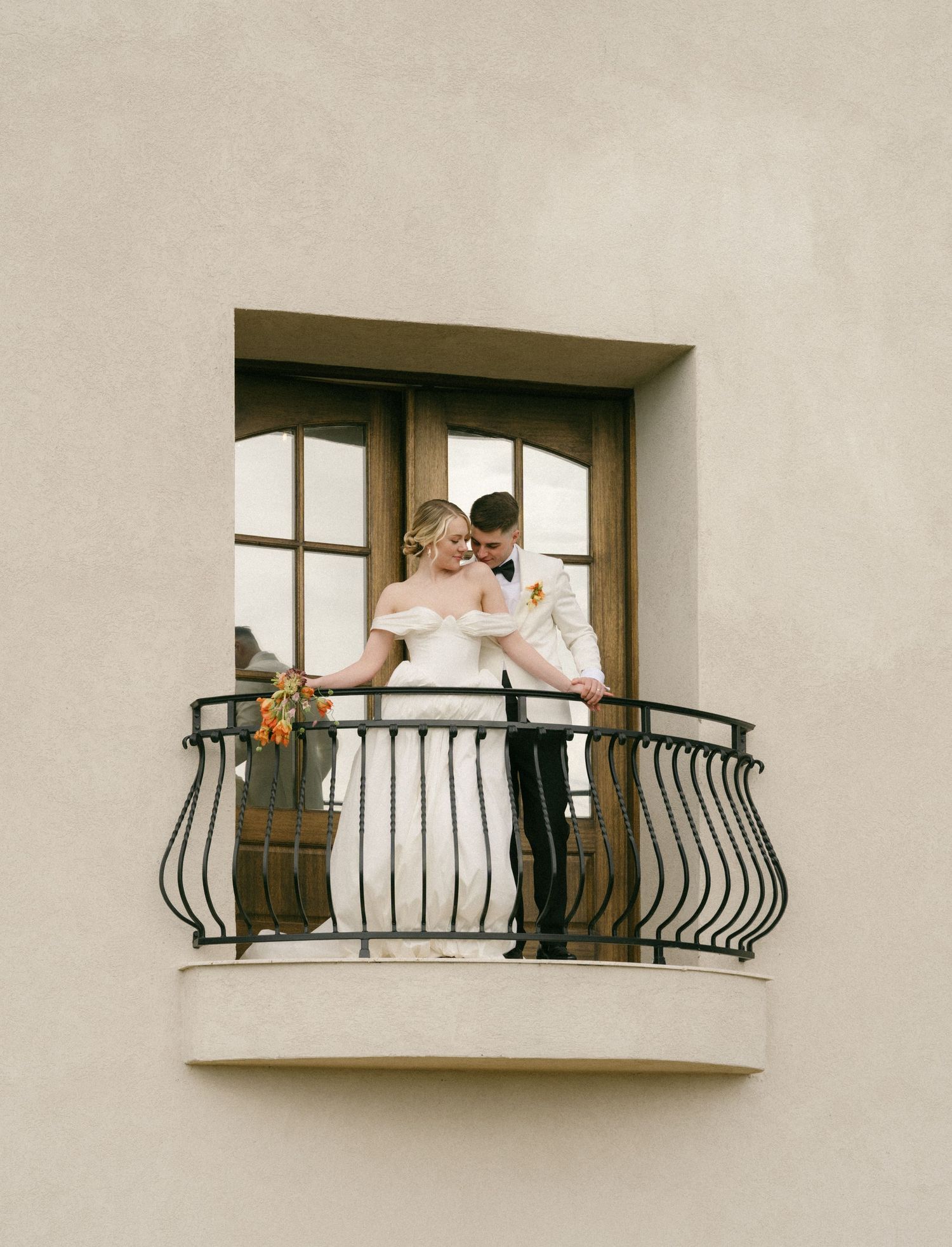 Bride and groom share a romantic moment on an elegant wrought iron balcony, bride holding a colorful bouquet.
