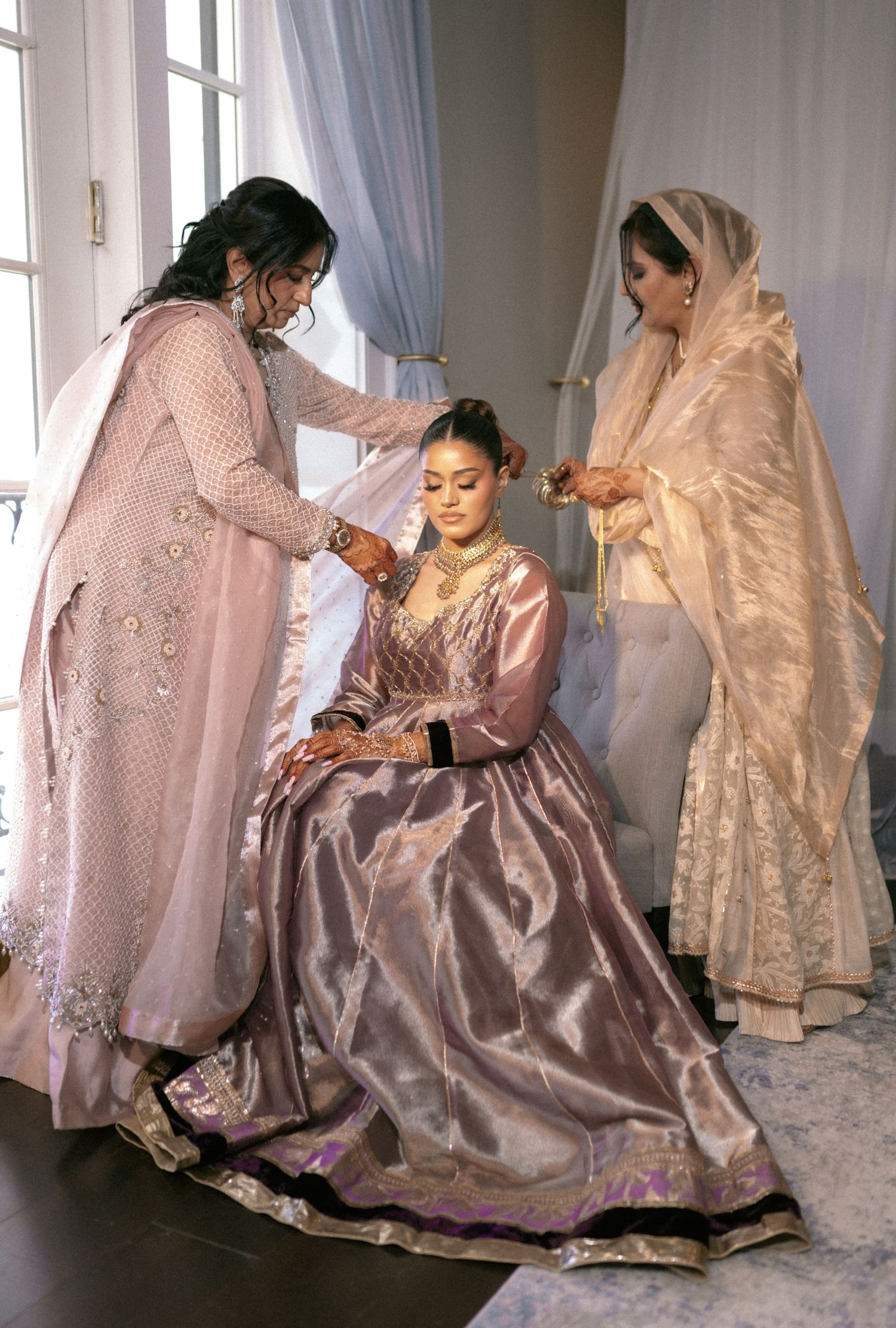 Bride in mauve lehenga being dressed by two women in traditional South Asian attire before wedding.