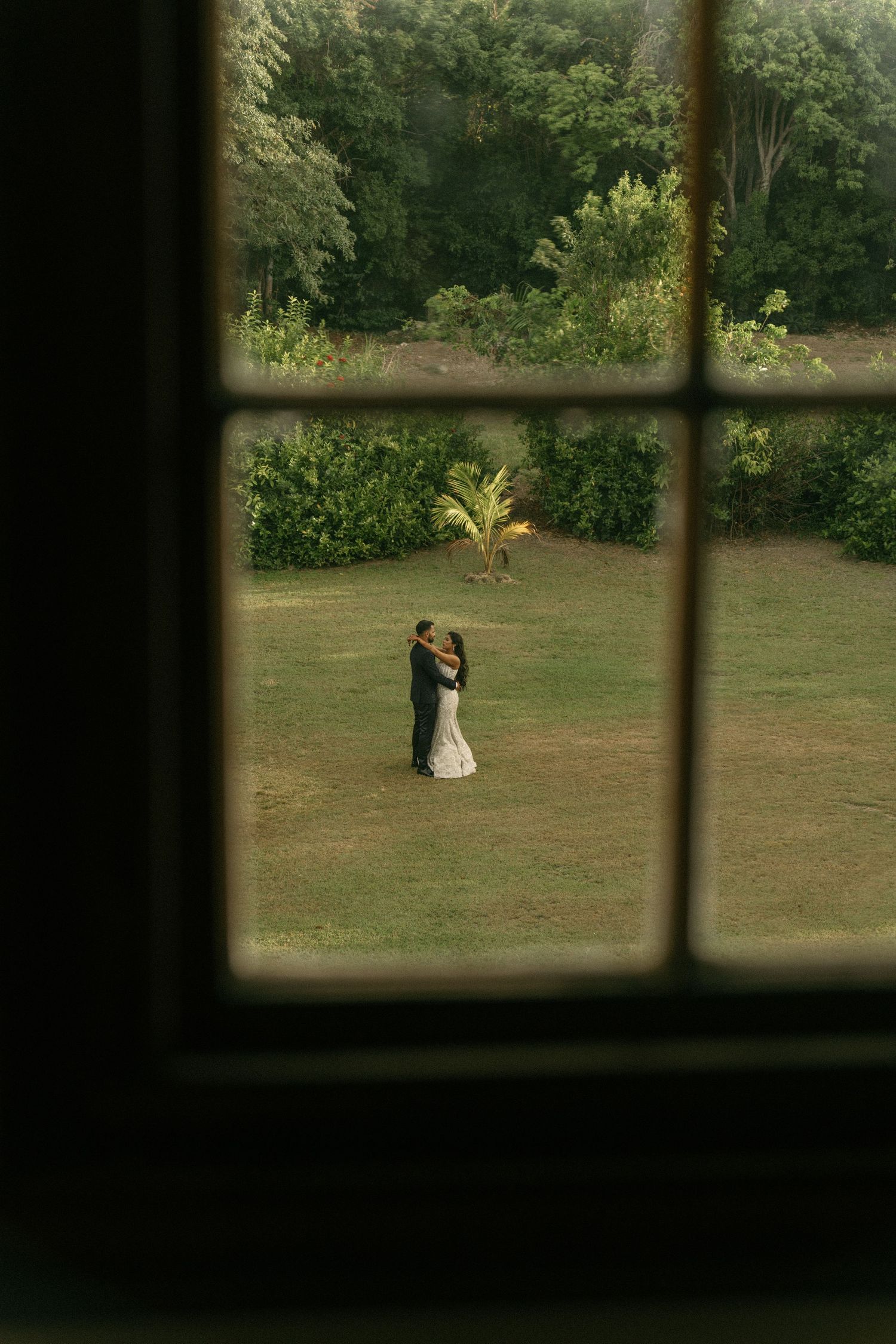 Bride and groom embrace on lush green lawn viewed through vintage window frame.