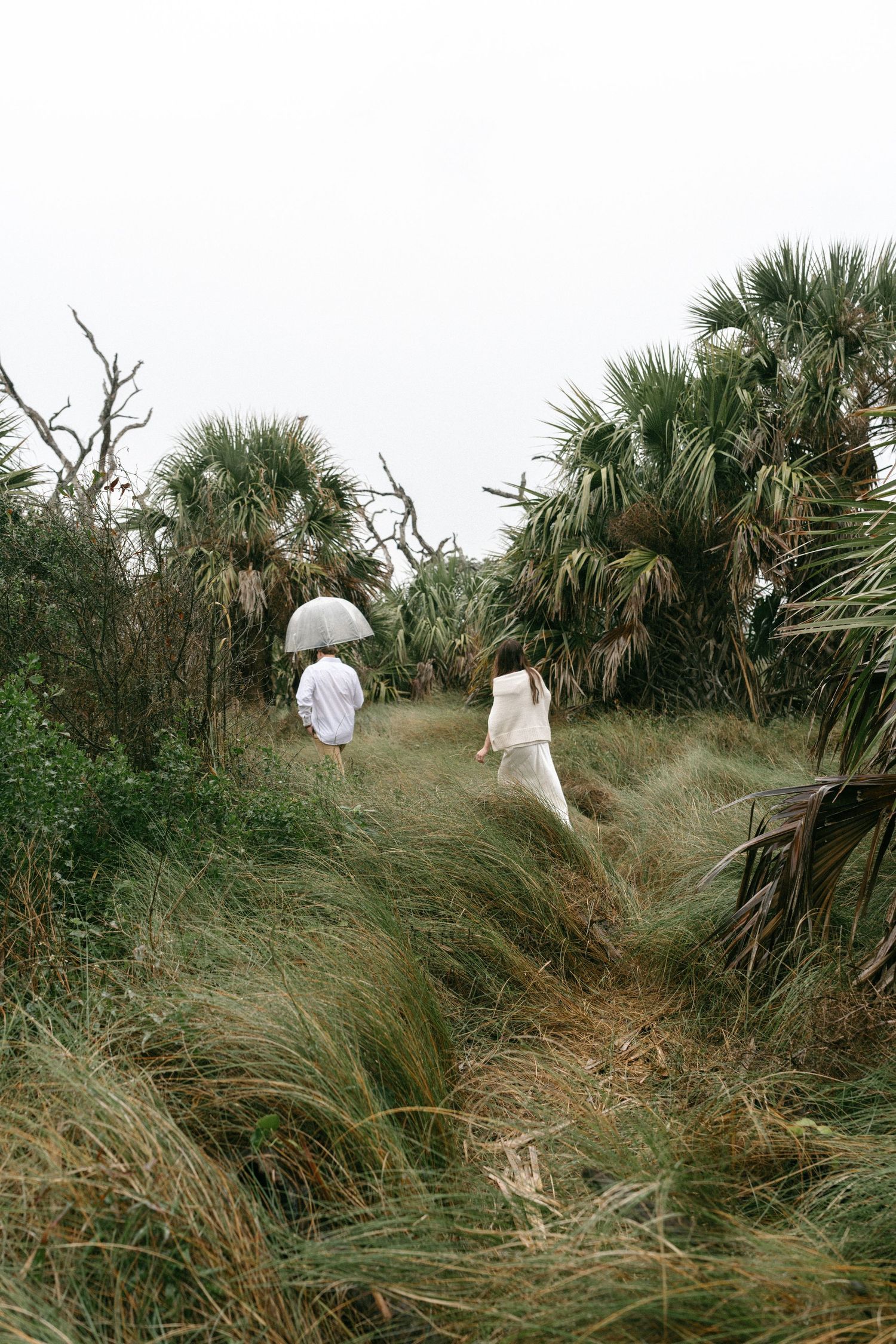 Two people in white attire walk through tall grass toward palm trees on a misty, overcast day.