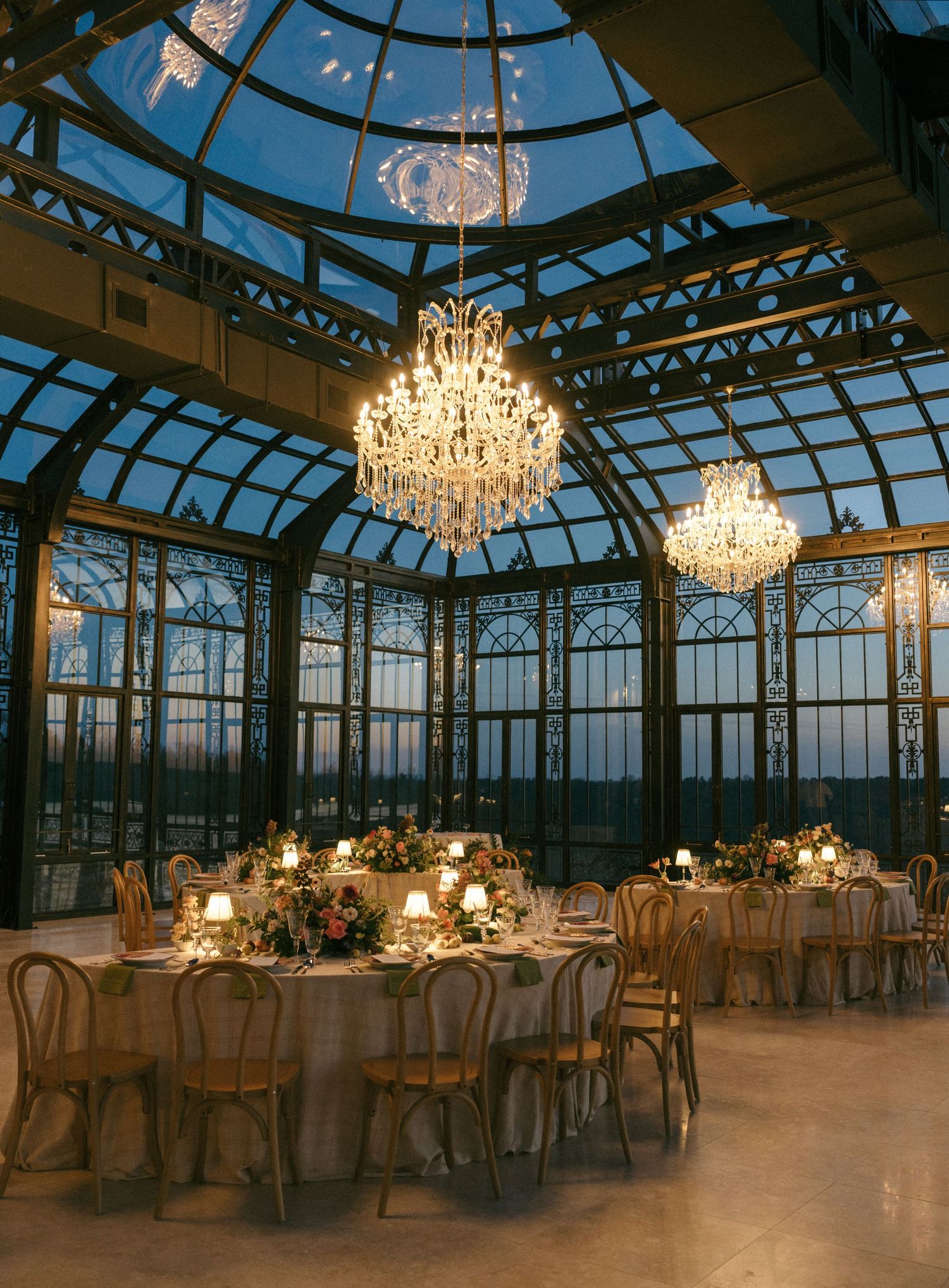 Elegant glass conservatory wedding reception with crystal chandeliers, candlelit round tables, and a stunning dusk sky backdrop.
