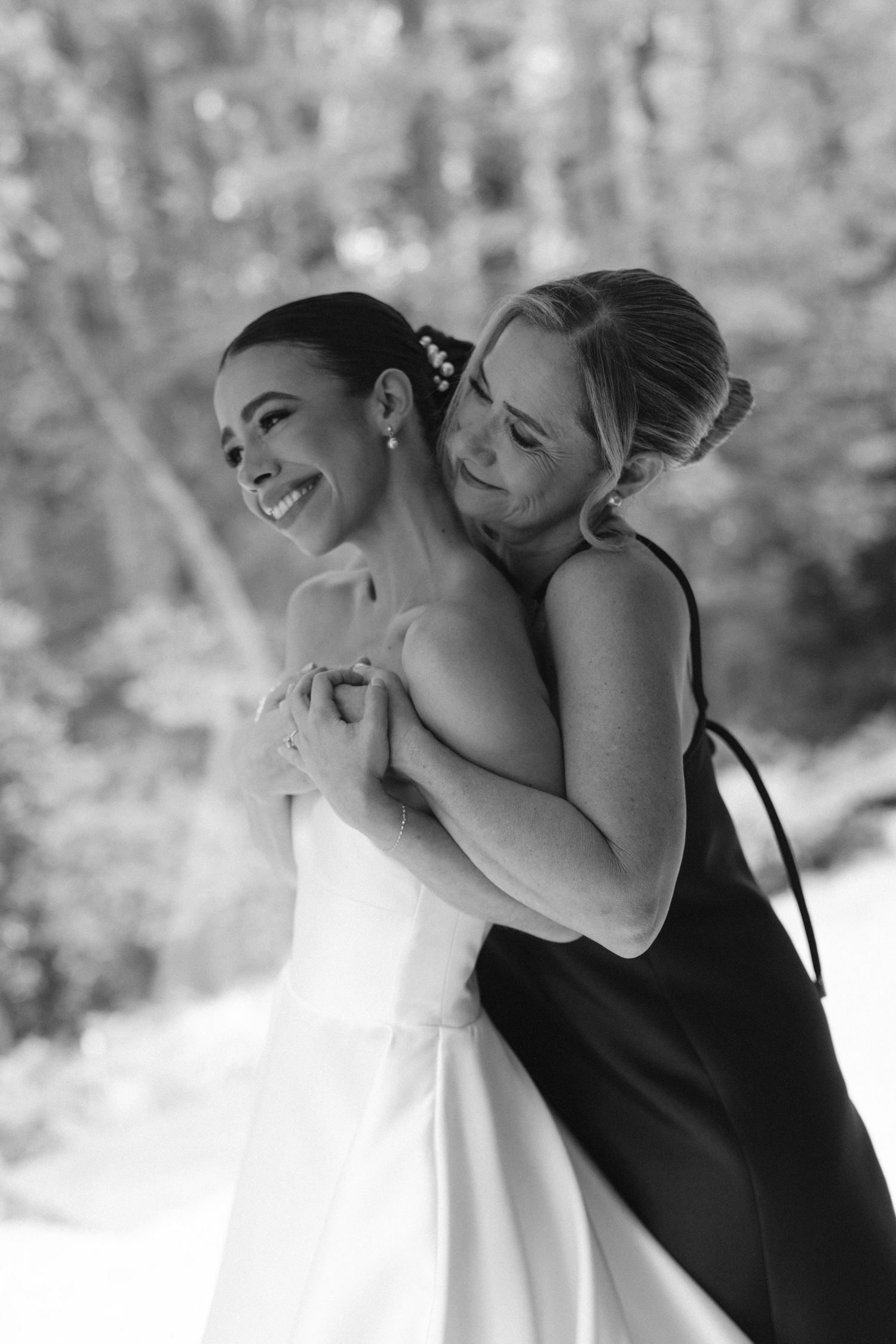 Black and white photo of bride in white gown being embraced from behind by bridesmaid in dark dress outdoors.