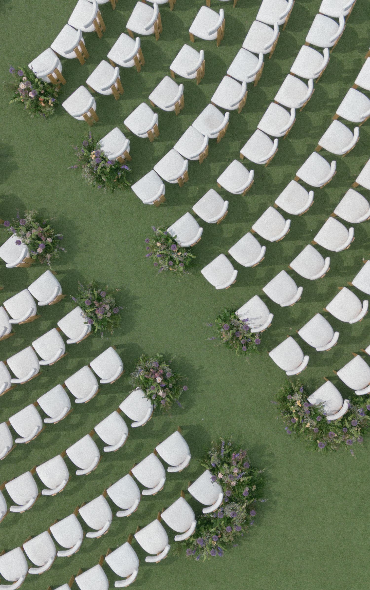 Aerial view of white chairs arranged in curved rows on green grass with floral arrangements along the aisle for an outdoor wedding.