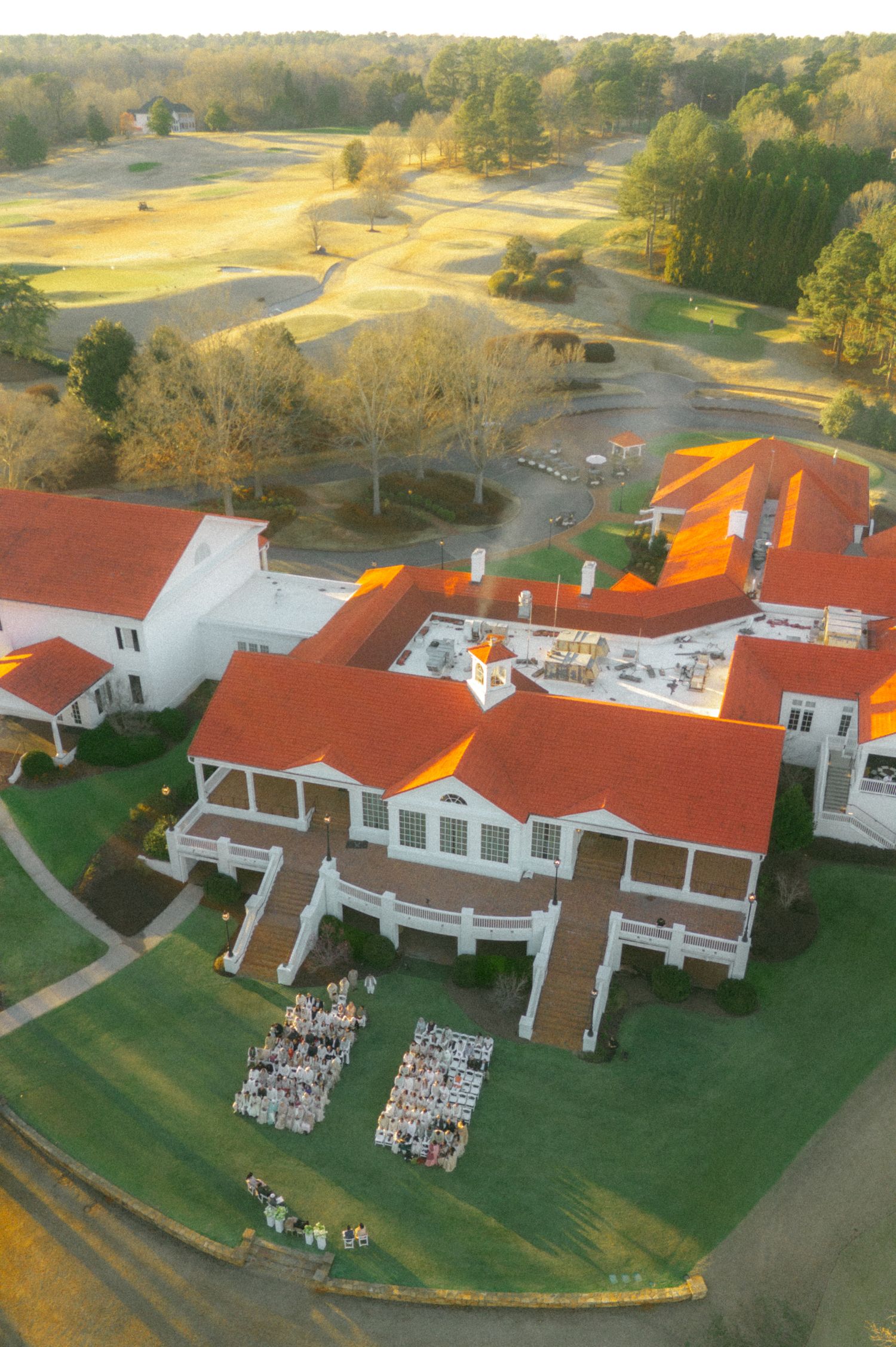 Aerial view of a elegant golf club with red roofs, white building, outdoor seating, and lush green fairways at sunset.