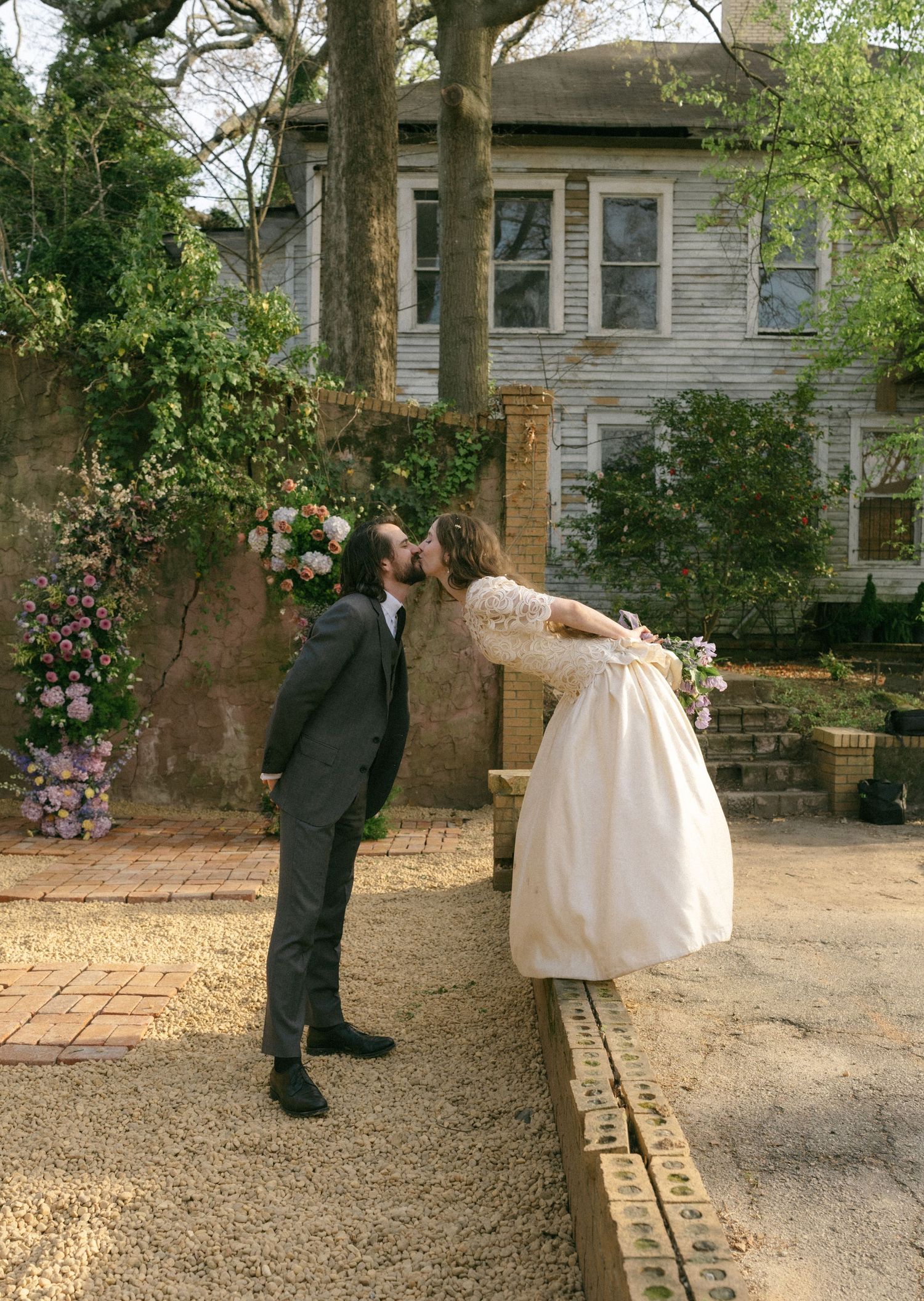Bride and groom sharing a kiss in a garden wedding venue with floral arch and historic home backdrop.