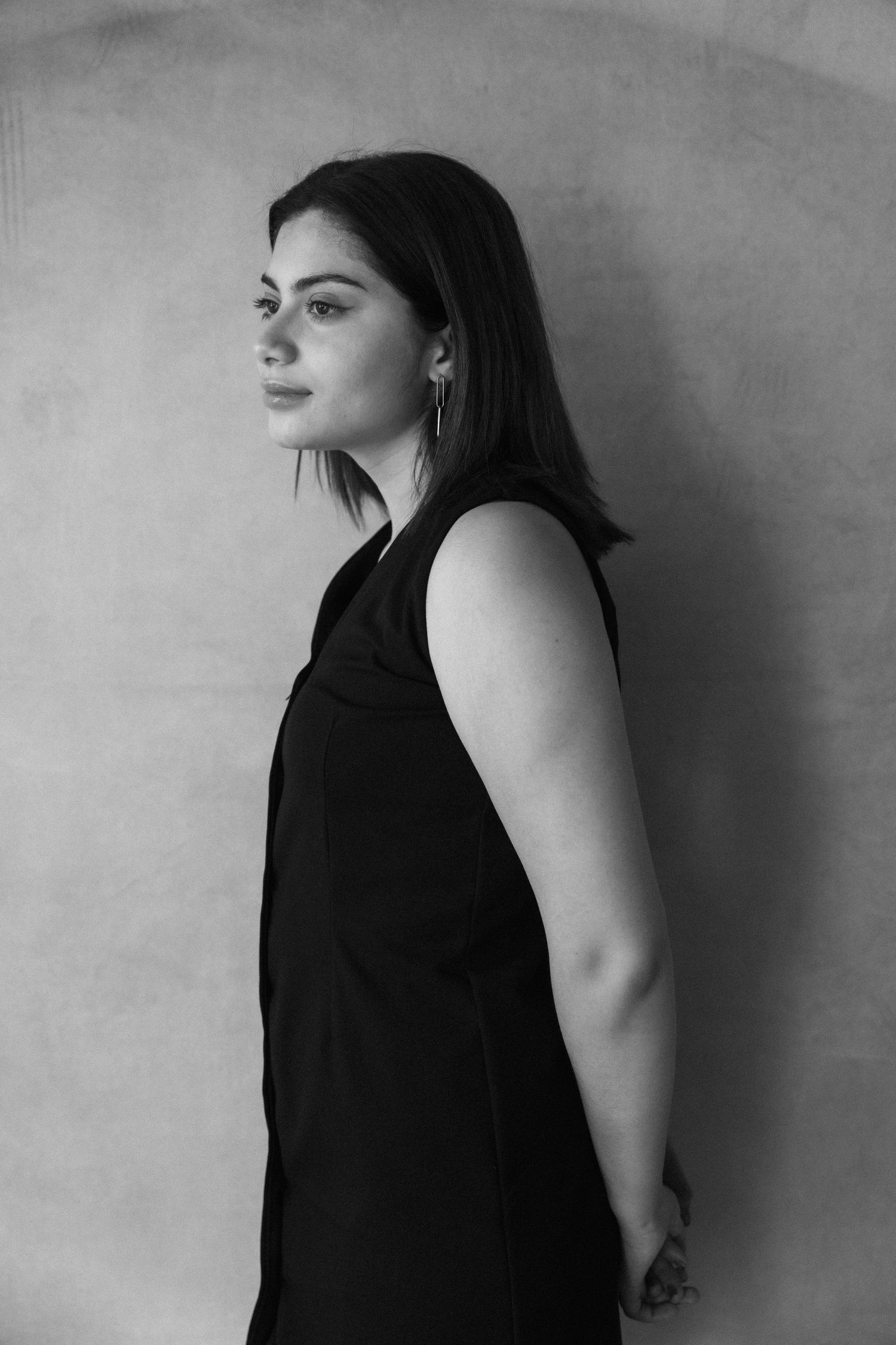 Black and white portrait of a young woman in a sleeveless black dress, gazing sideways against a neutral background.