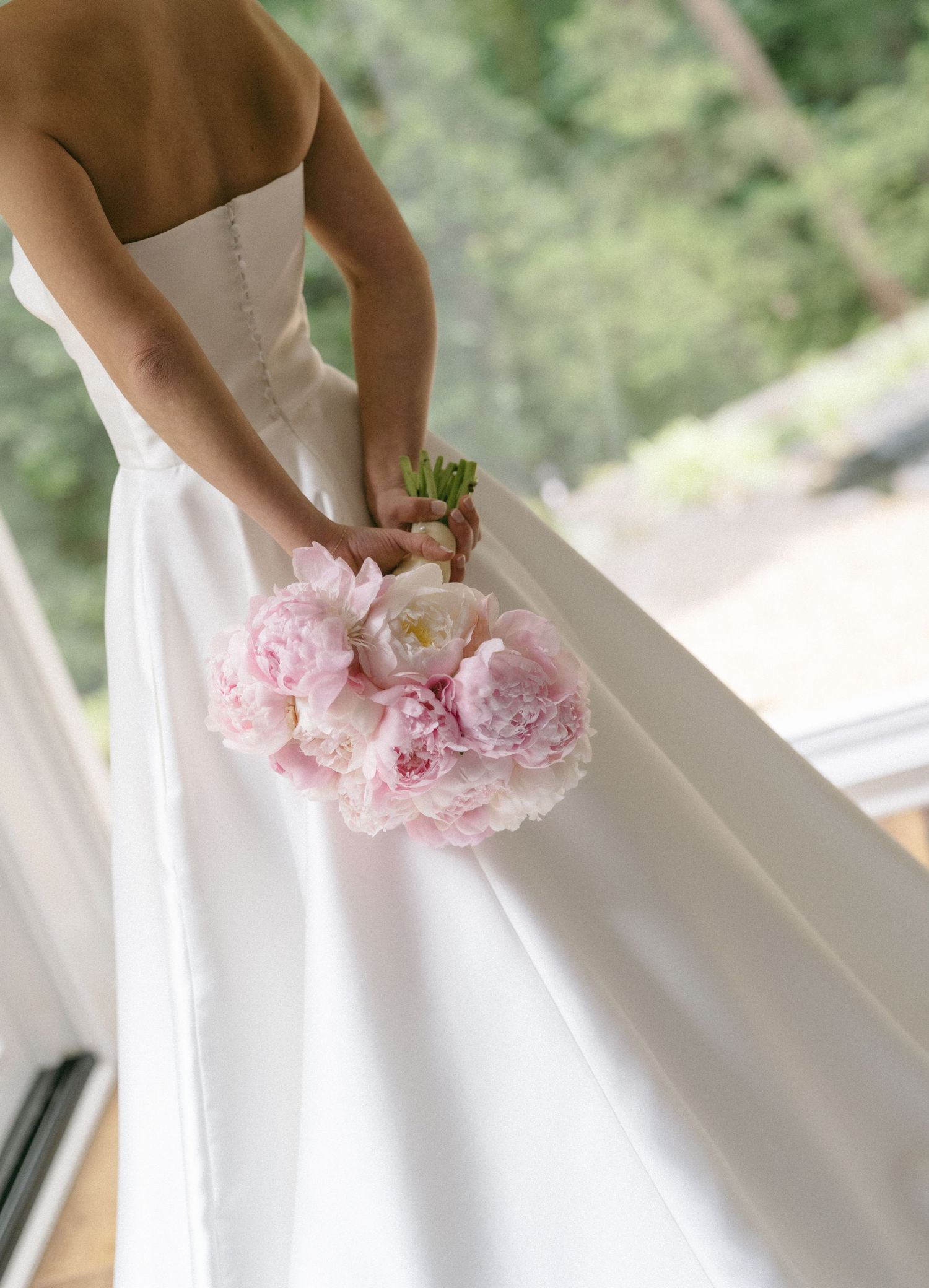 Bride in strapless white gown holding a lush pink peony bouquet near a sunlit window.