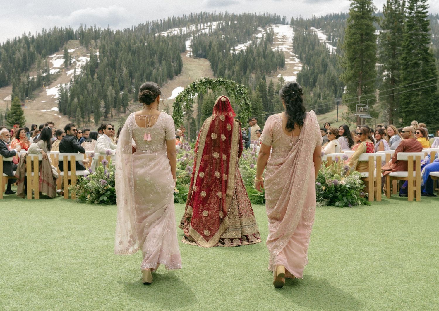 Indian bride in red lehenga walks with two bridesmaids in pink sarees toward outdoor mountain wedding ceremony.