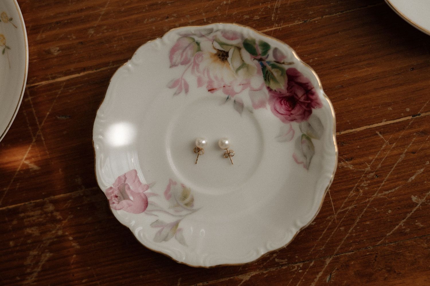 Vintage white porcelain saucer with pink rose pattern holding a pair of small pearl stud earrings on a wooden surface.