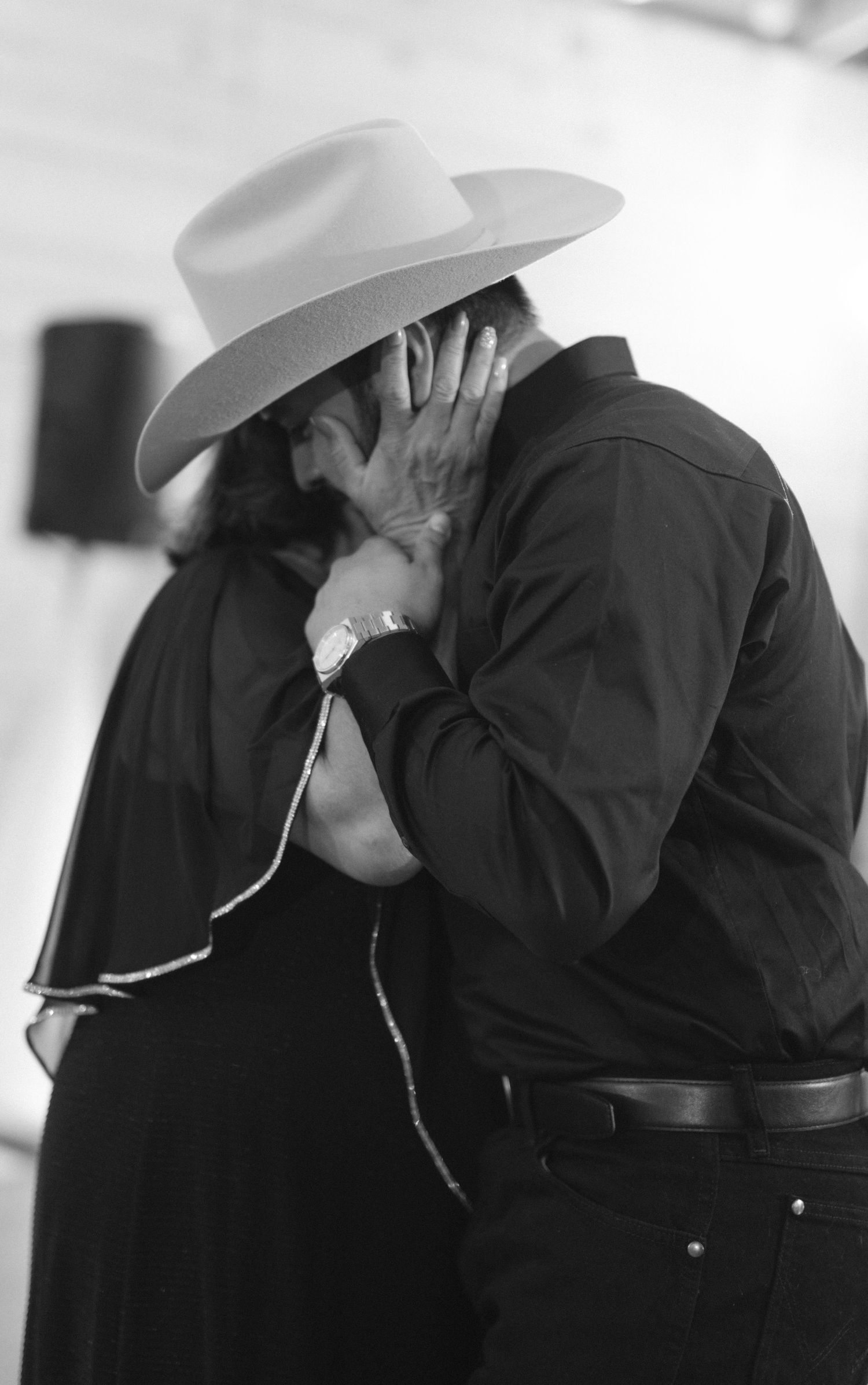 Black and white photo of a couple sharing a romantic kiss, man wearing a cowboy hat and watch, dressed in western attire.