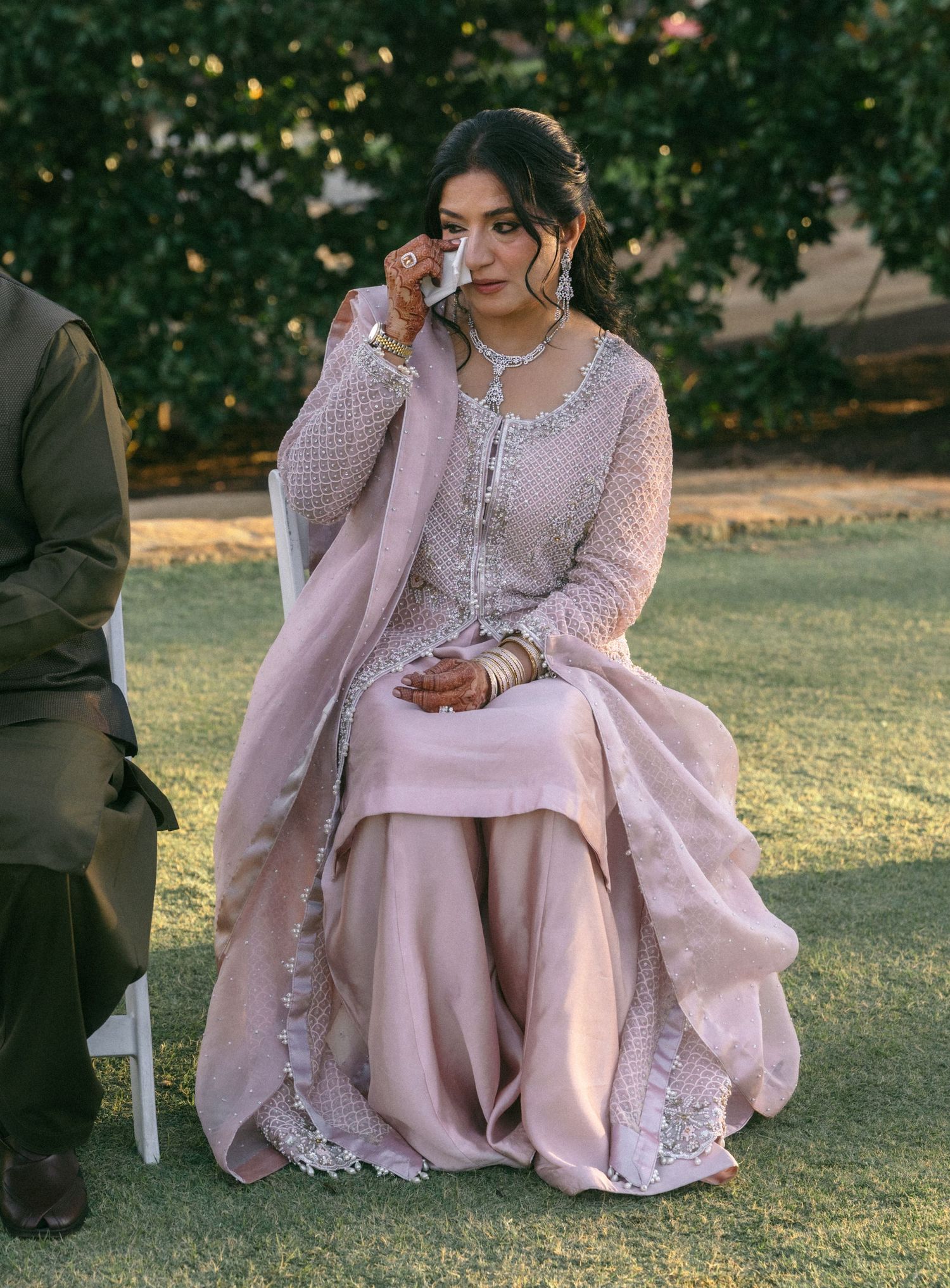 Woman in pink embroidered salwar kameez wiping tears while seated outdoors at a formal South Asian event.