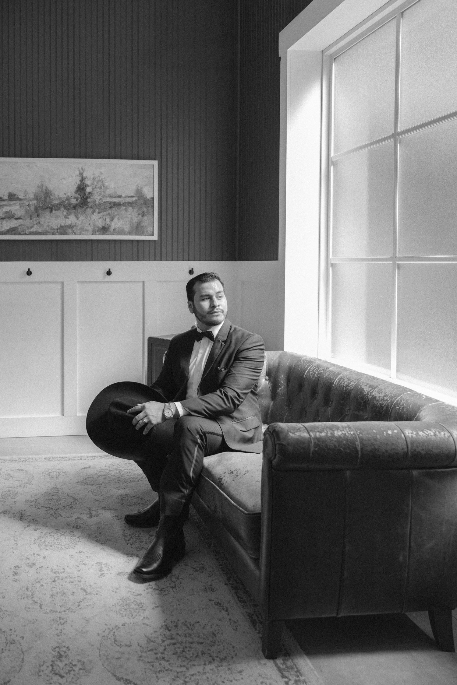 Elegant man in suit holding hat seated on tufted leather sofa in black and white portrait photo.