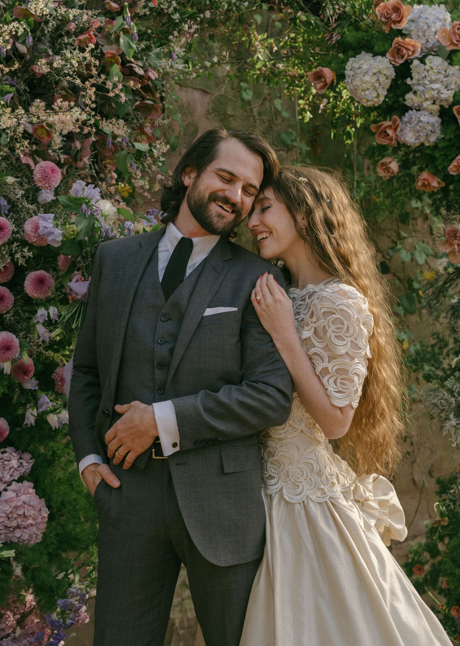 A smiling couple in wedding attire poses romantically before a lush floral backdrop of pink and white blooms.