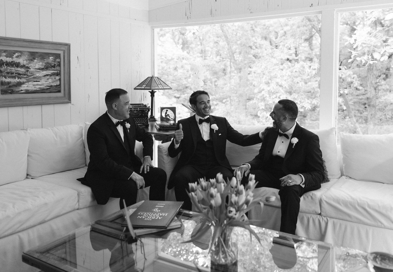 Three groomsmen in tuxedos celebrate together in a bright white room during a wedding day preparation.