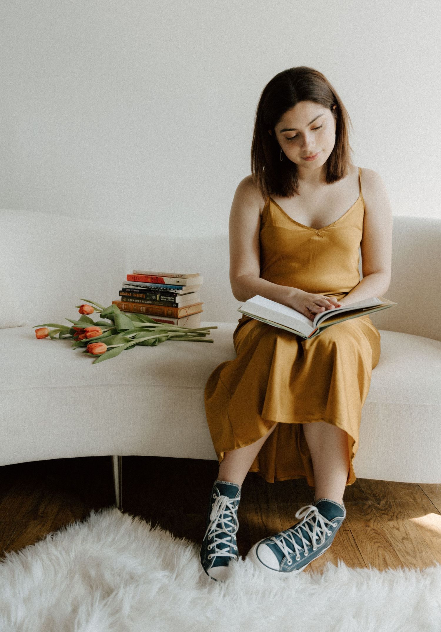 Woman in gold dress reading a book on white sofa with tulips and stacked books beside her.