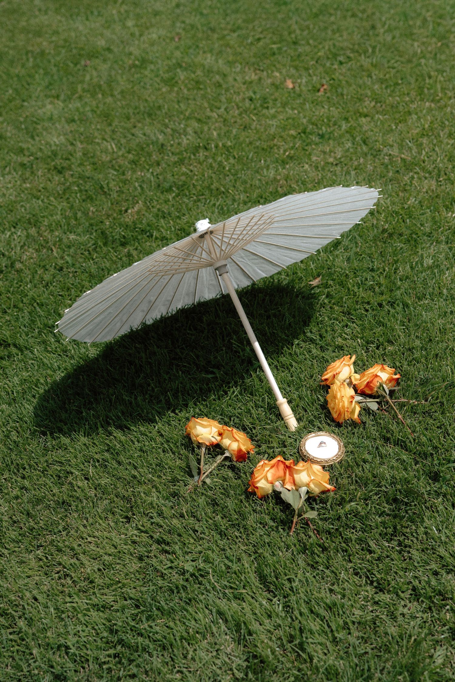 A white paper parasol umbrella resting on green grass beside orange roses and a candle.