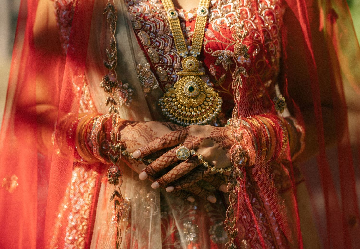 Indian bride in red lehenga with gold jewelry and mehndi-adorned hands clasped together.
