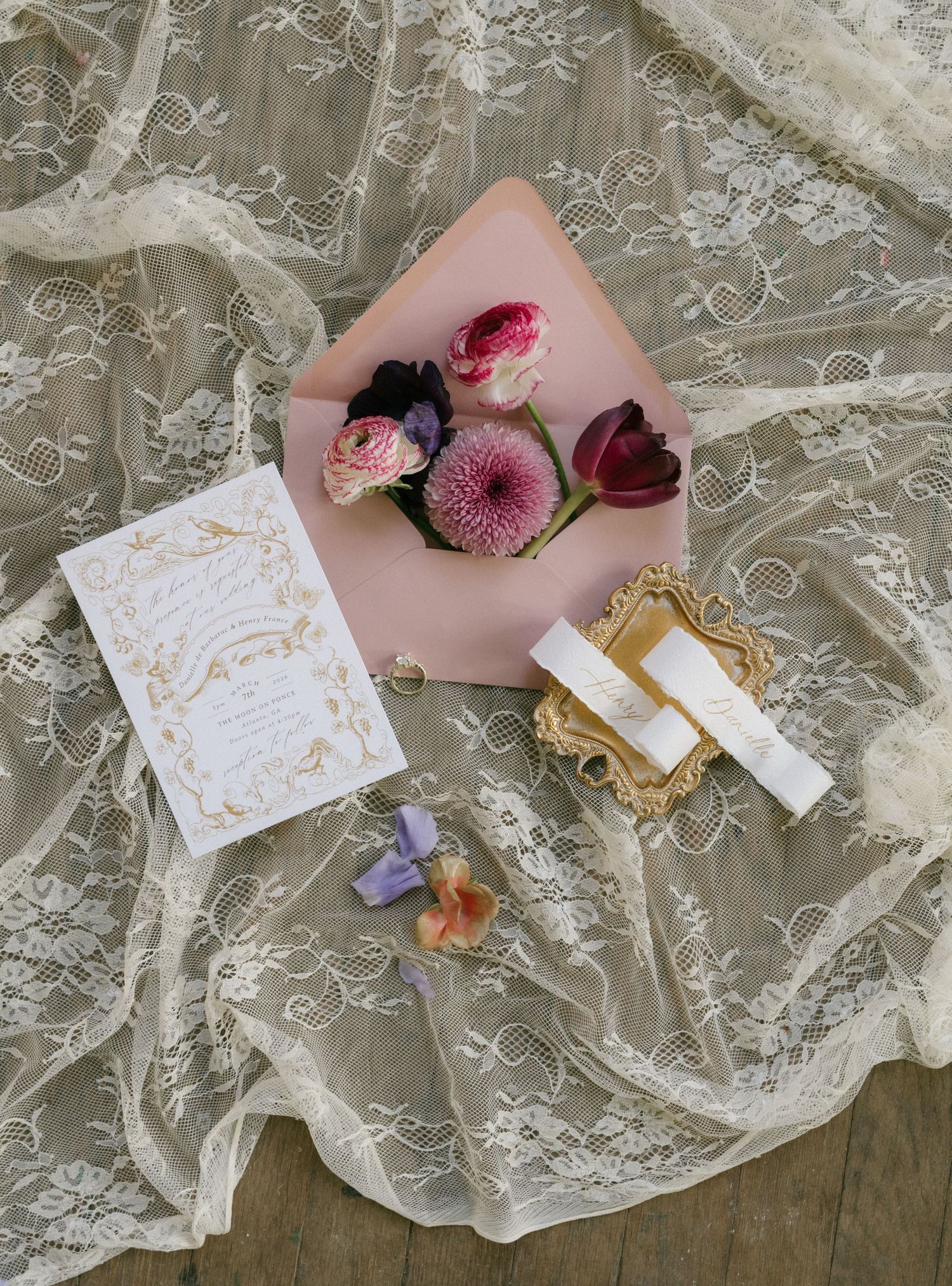 Romantic wedding flat lay with gold calligraphy invitation, pink envelope, fresh flowers on delicate lace fabric.