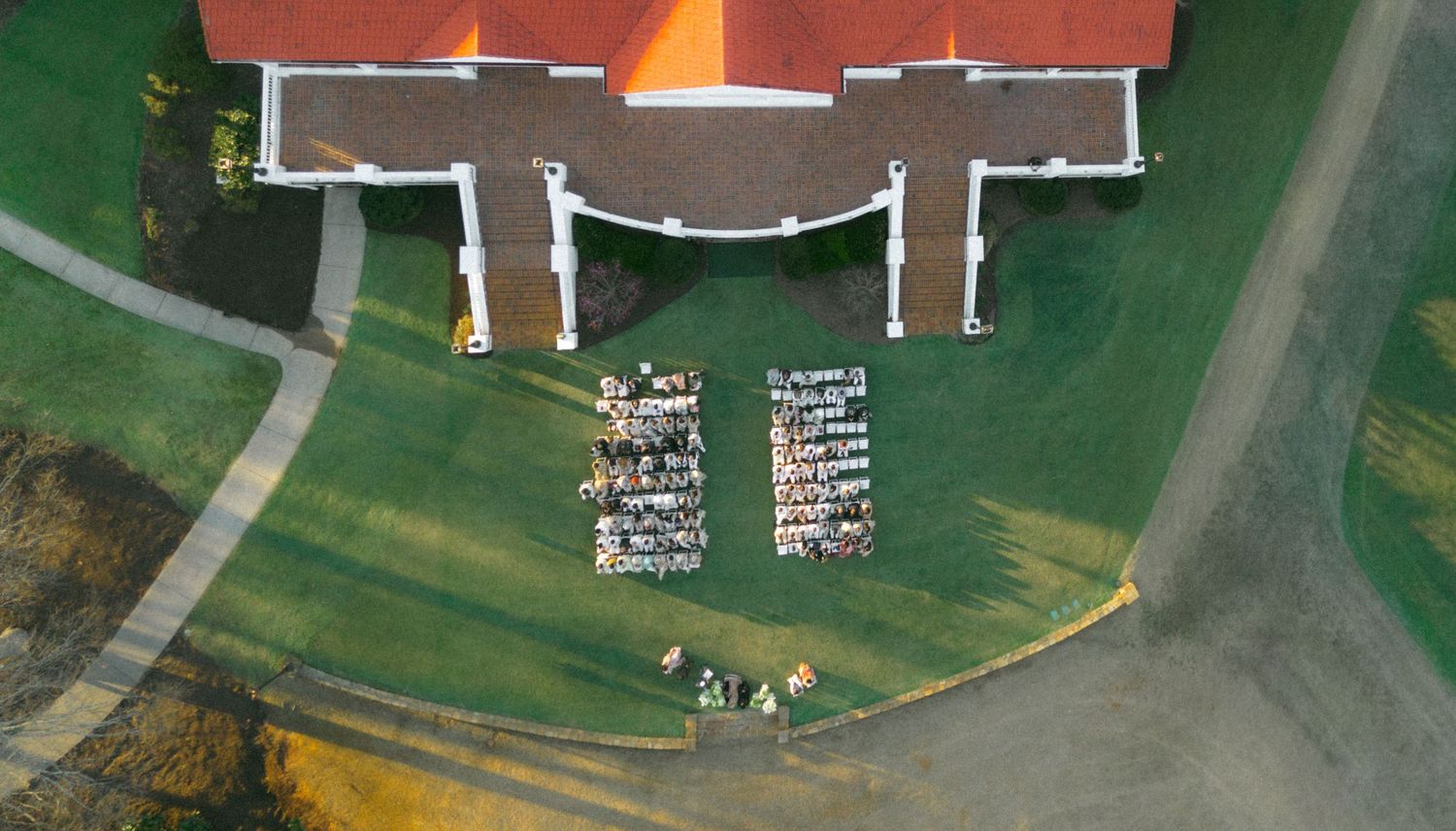 Aerial view of a red-roofed building with white columns, surrounded by green lawns and two groups of seated wedding guests.