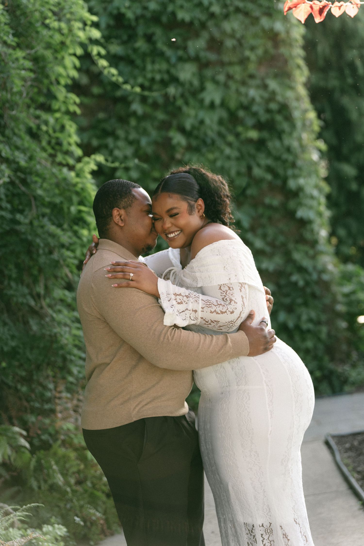 Happy couple embracing and smiling in front of a lush green ivy-covered wall outdoors.