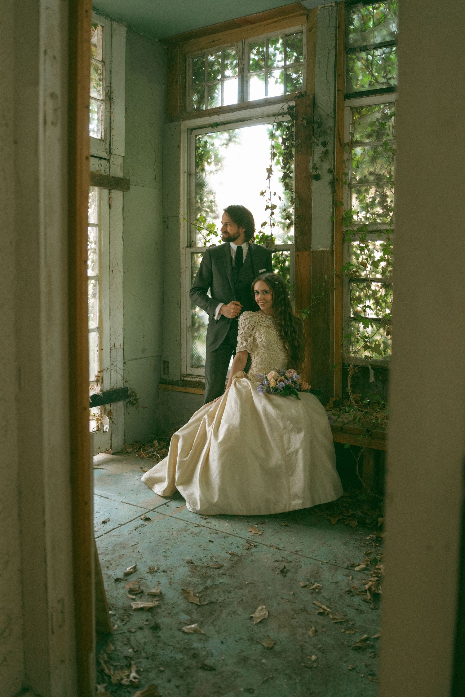 Bride and groom posing in a rustic abandoned room with ivy-covered windows and vintage charm.