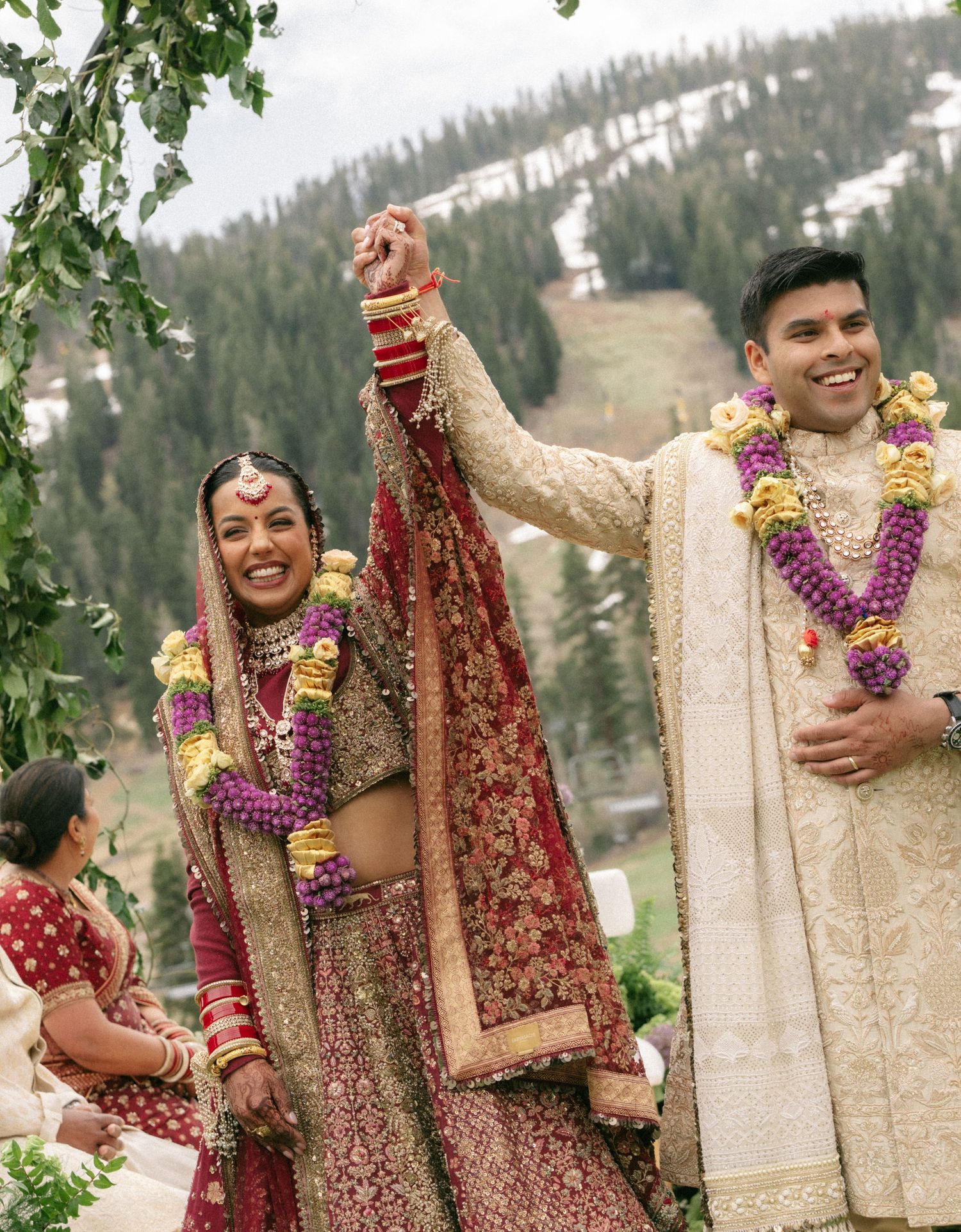 Indian bride and groom in traditional attire celebrate at outdoor wedding ceremony with mountain backdrop.