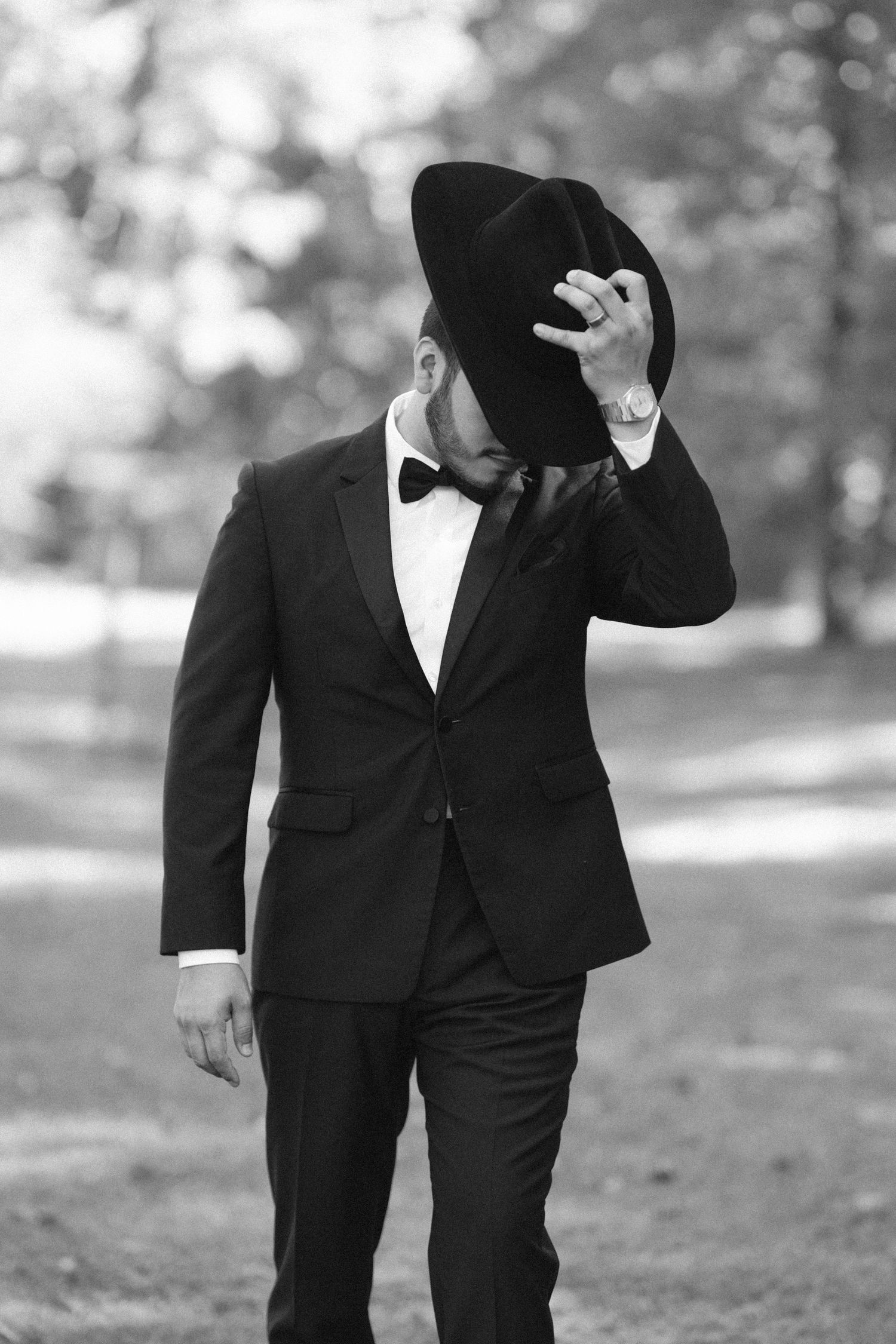 Black and white photo of a dapper man in a tuxedo and bow tie tipping his wide-brimmed hat in a park.