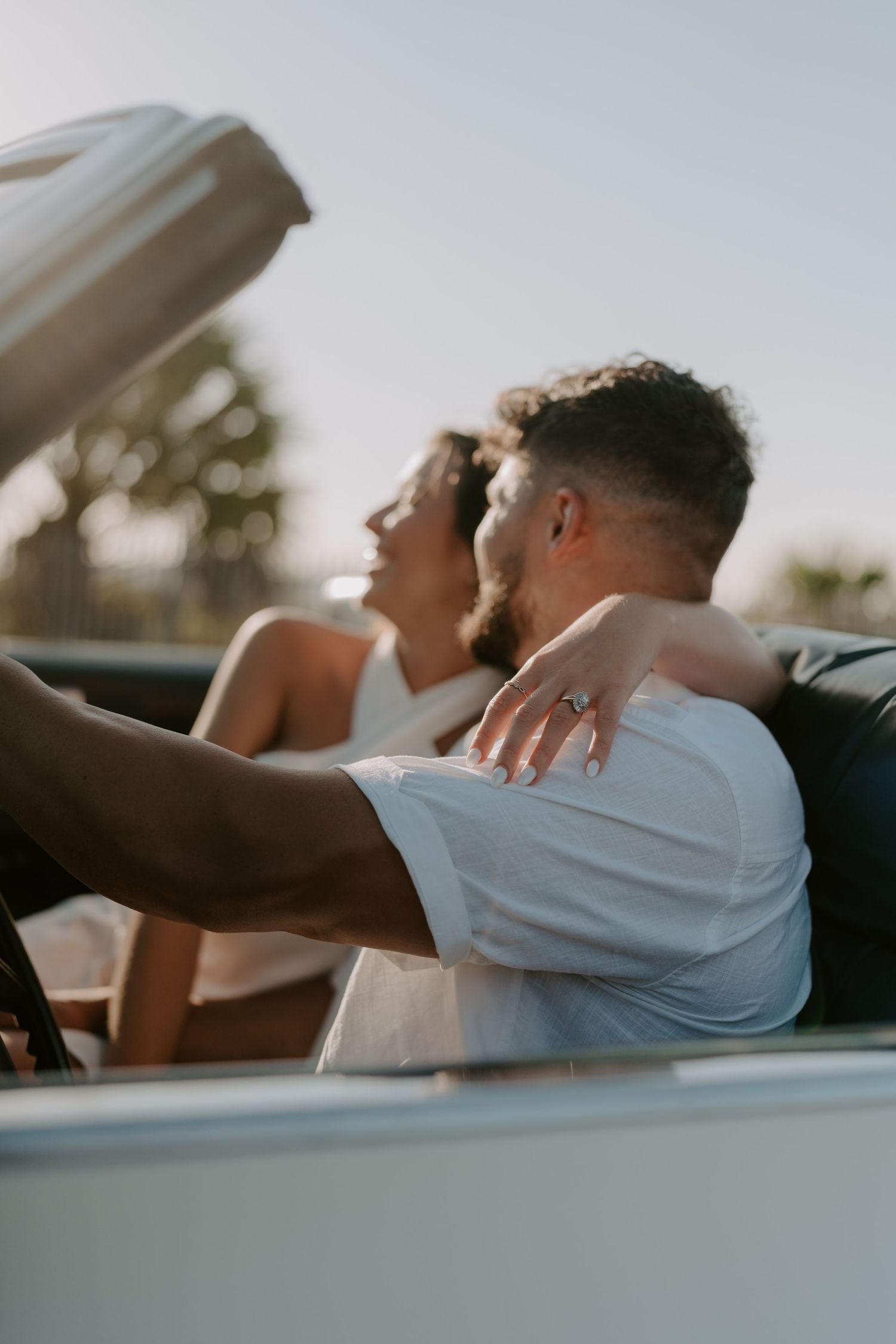 Couple sitting in a convertible car, woman with ring on hand embracing man, golden hour sunlight in background.