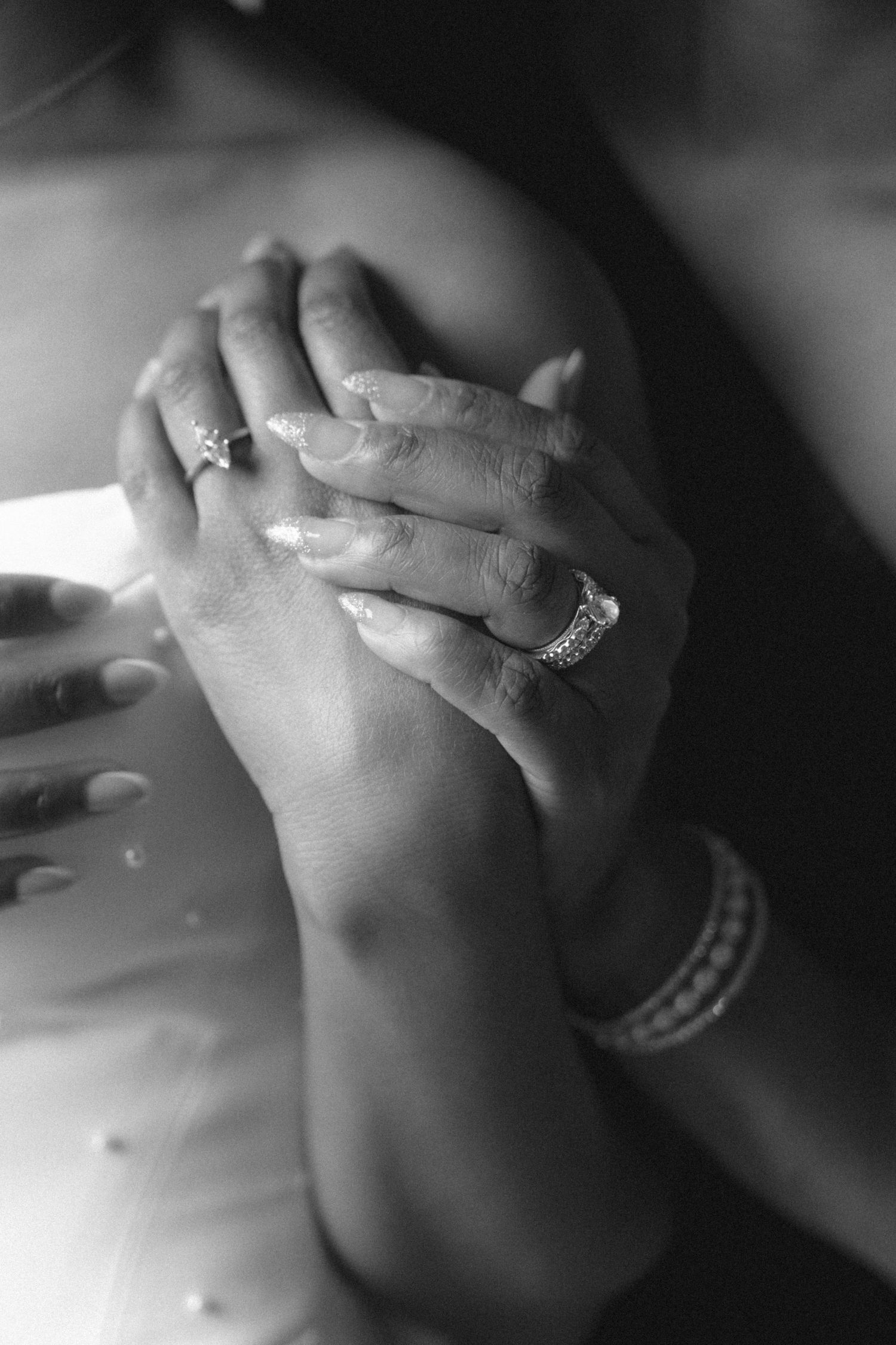 Close-up of bride and groom holding hands, showcasing wedding rings and bracelet in black and white.