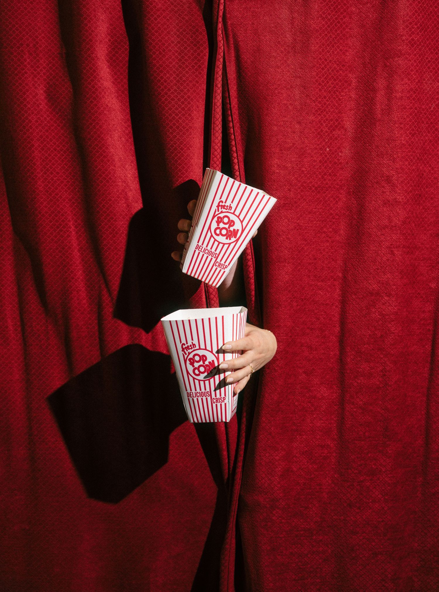 Two hands reaching through red curtains holding classic striped popcorn boxes at a movie theater.