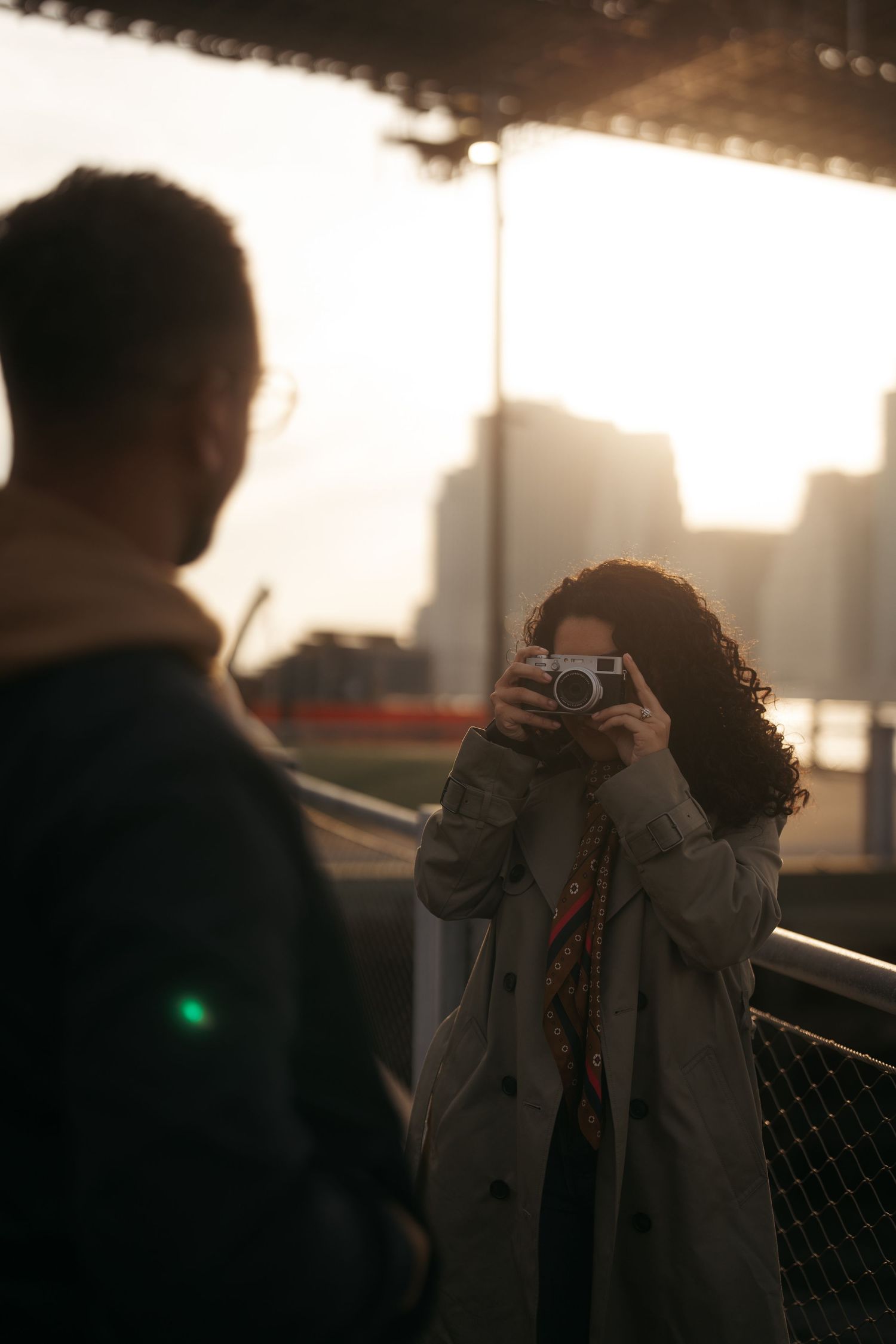 Woman photographing man with film camera at golden hour on urban bridge.