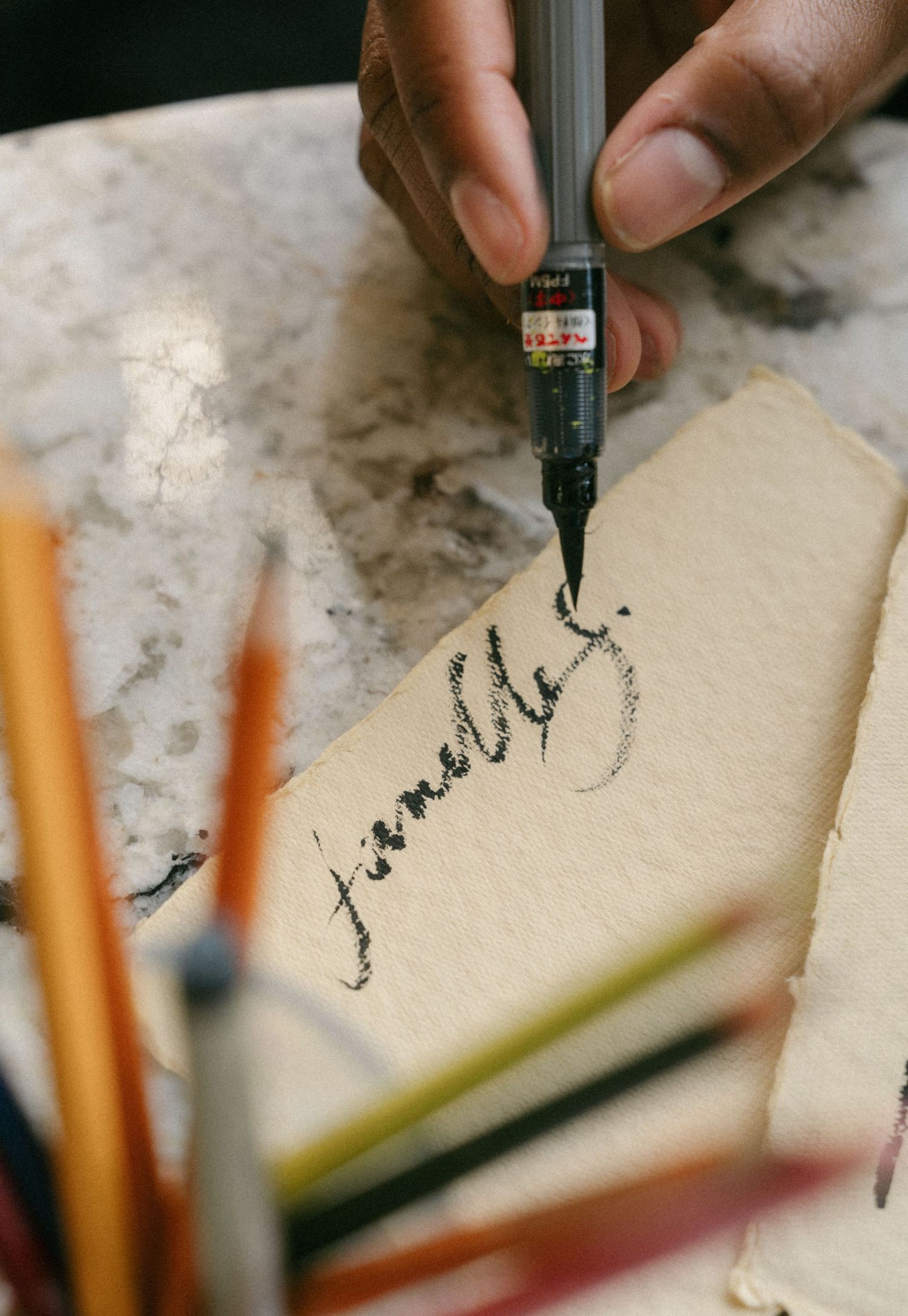 A hand writes cursive calligraphy on cream paper using a black ink brush pen on a marble surface.