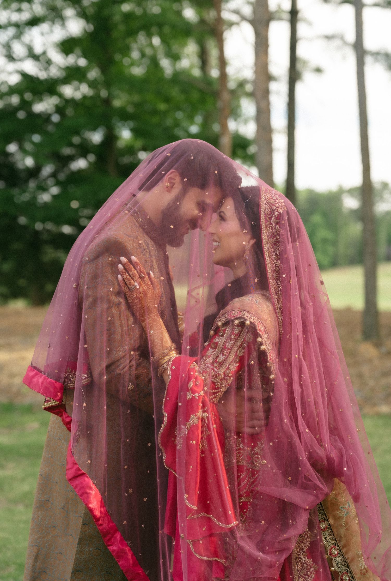 South Asian couple sharing romantic moment under pink dupatta veil outdoors at wedding ceremony.