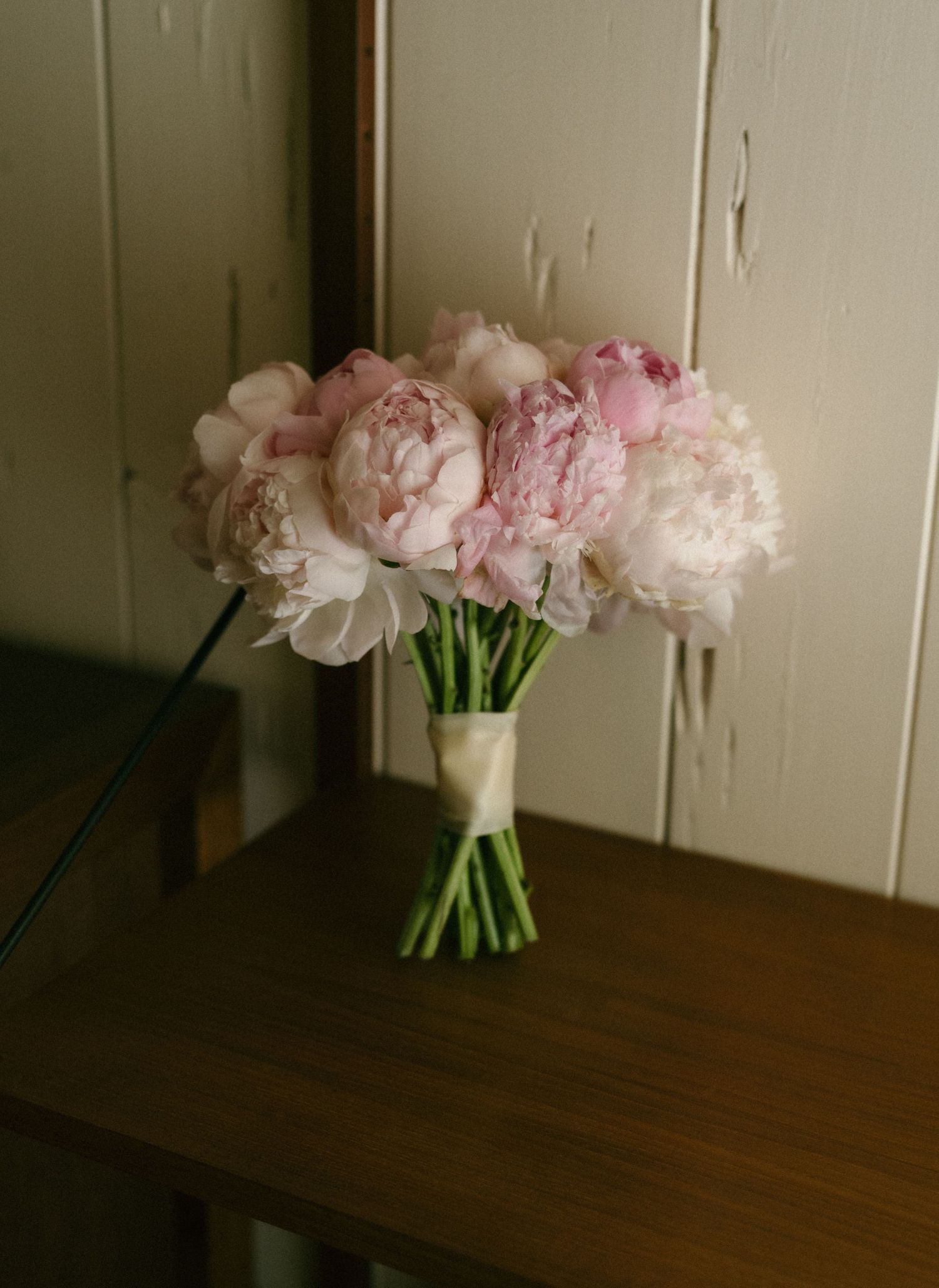 Pink and white peony bridal bouquet wrapped with ivory ribbon resting on a wooden table.