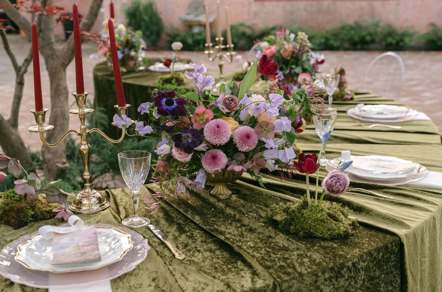 Elegant outdoor tablescape with green velvet runner, floral centerpiece, red taper candles, and gold accents.