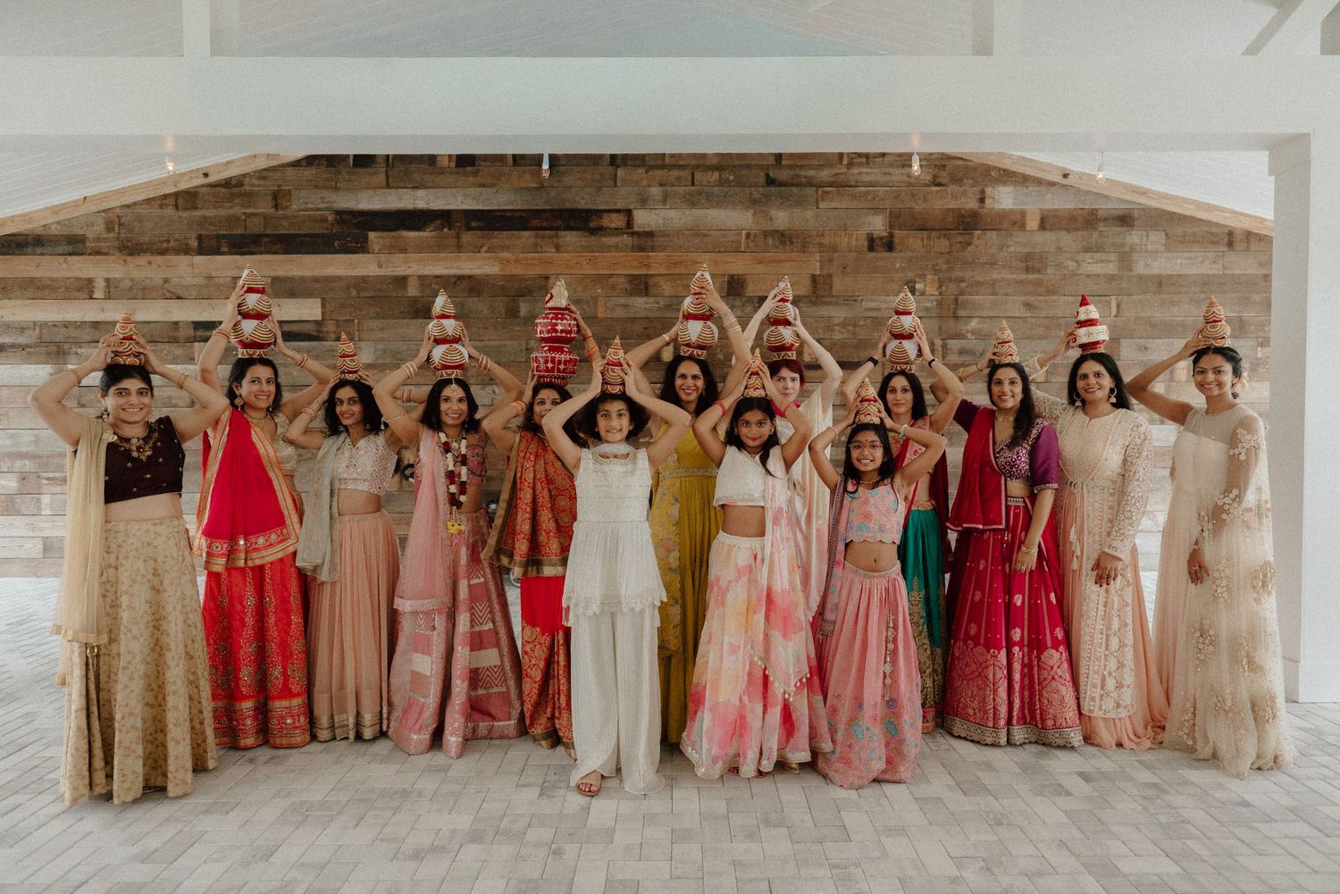 A group of women in colorful Indian bridal attire posing with arms raised at an elegant wedding celebration.