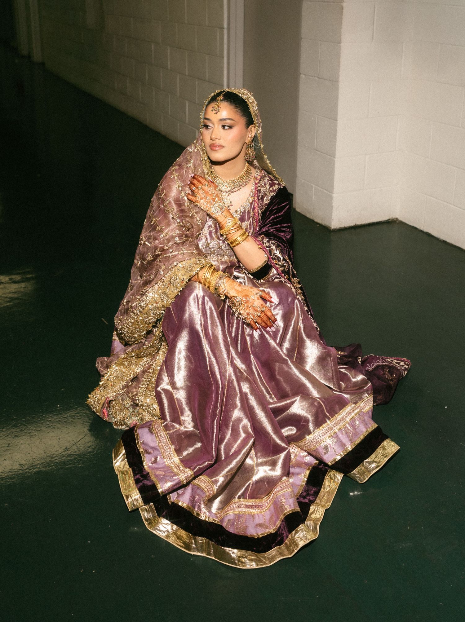 Woman in purple and gold bridal lehenga with dupatta, mehndi, and jewelry, seated gracefully on floor.
